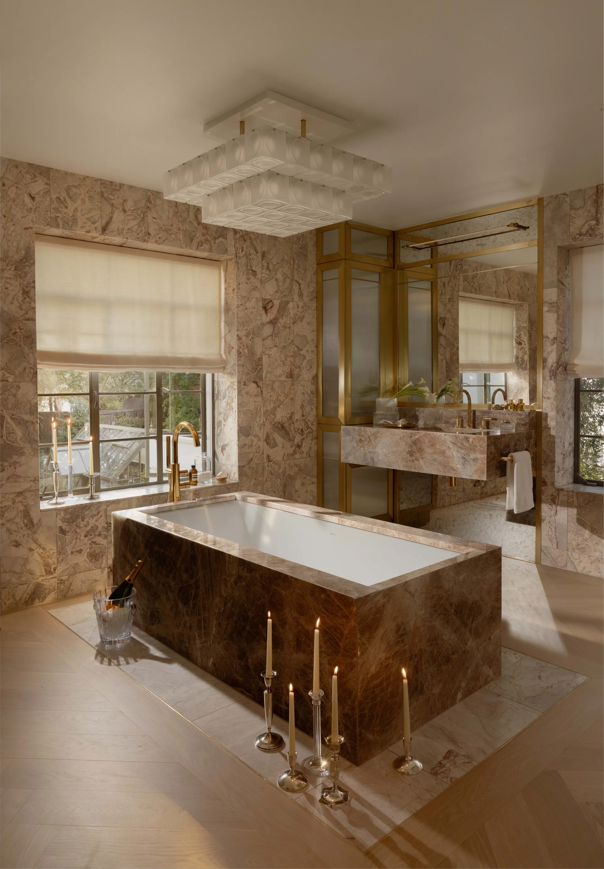 A bathroom featuring a large rectangular brown marble bathtub, matching marble walls, a floating sink vanity, and light herringbone wood floors.