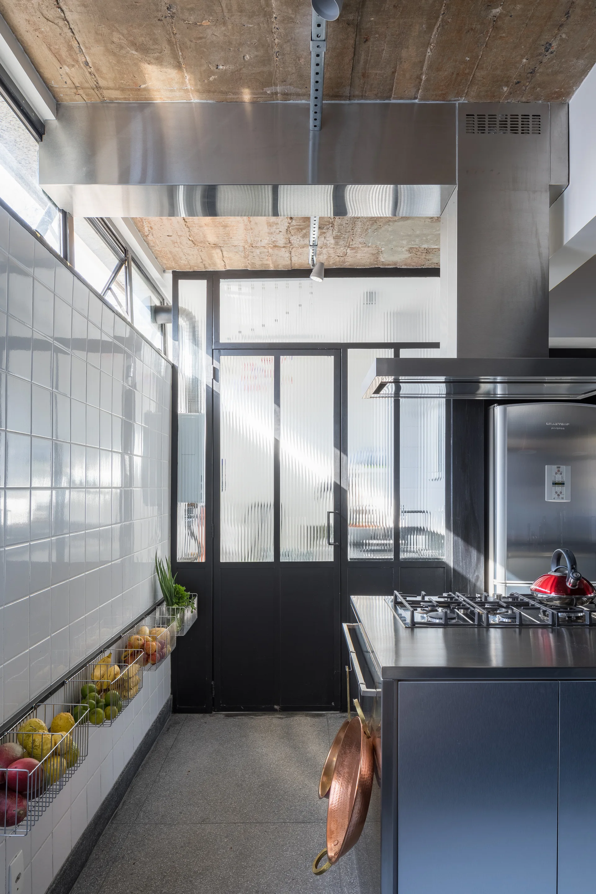 An industrial kitchen features a stainless steel island, white tiled walls, wire fruit baskets, and a fluted glass door beneath a concrete ceiling.