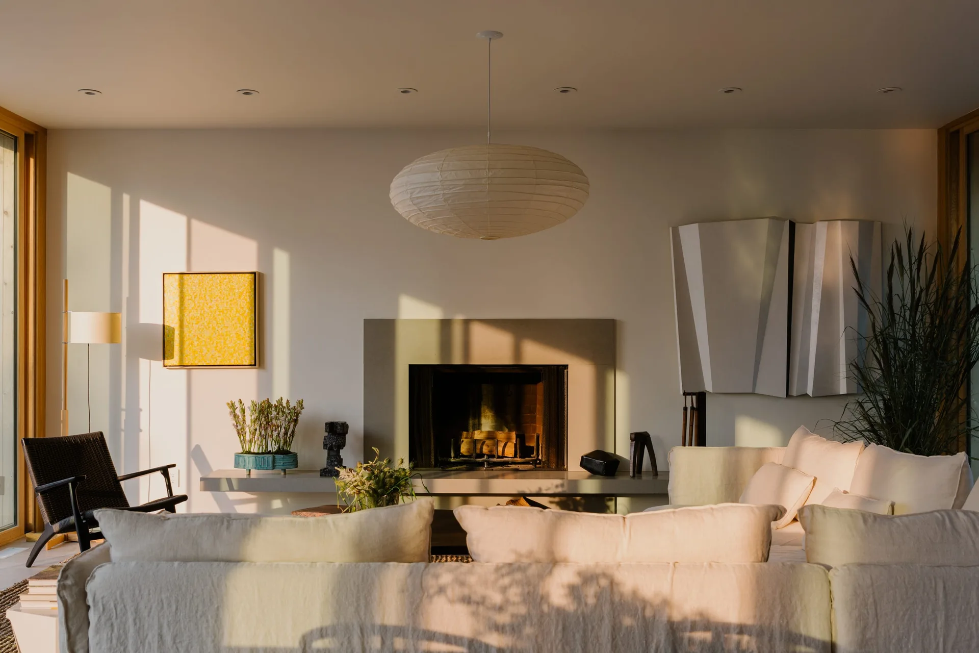 A sunlit minimalist living room featuring an off-white sectional sofa, a stone fireplace with an extended hearth, and a paper pendant light.