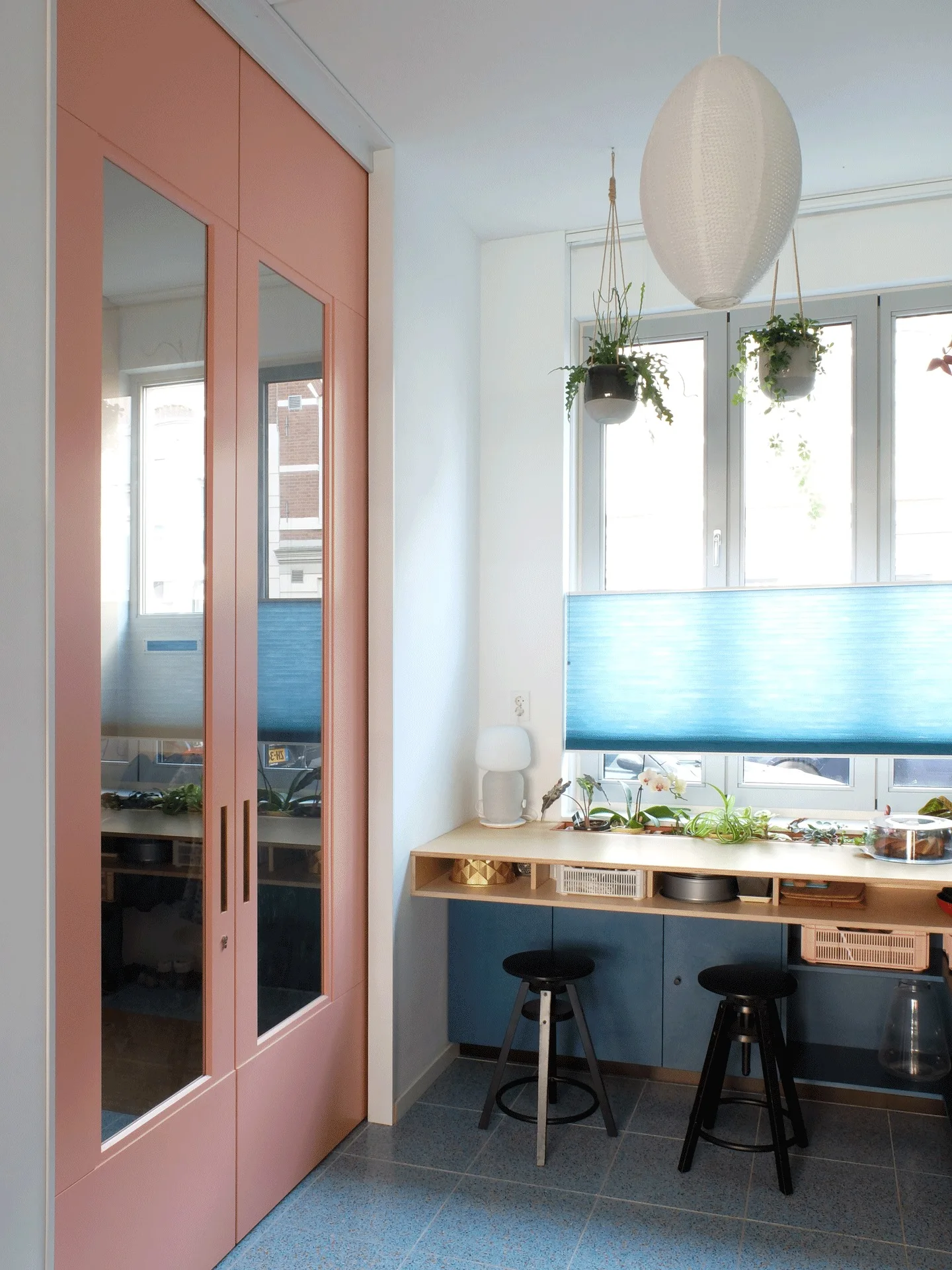 A tall pink storage cabinet with mirrored doors stands next to a wooden built-in desk with two black stools under a window with a blue shade.