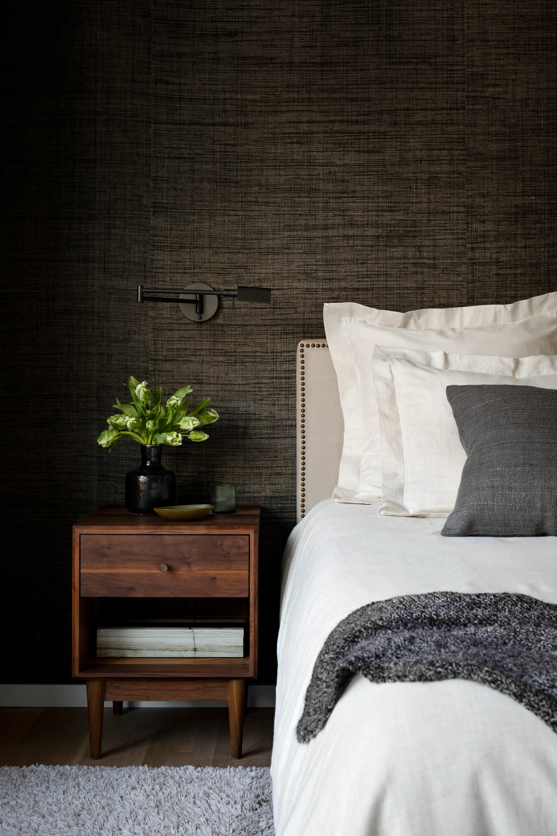 A beige headboard with nailhead trim sits against dark textured wallpaper next to a wood nightstand with white flowers and a swing-arm sconce.
