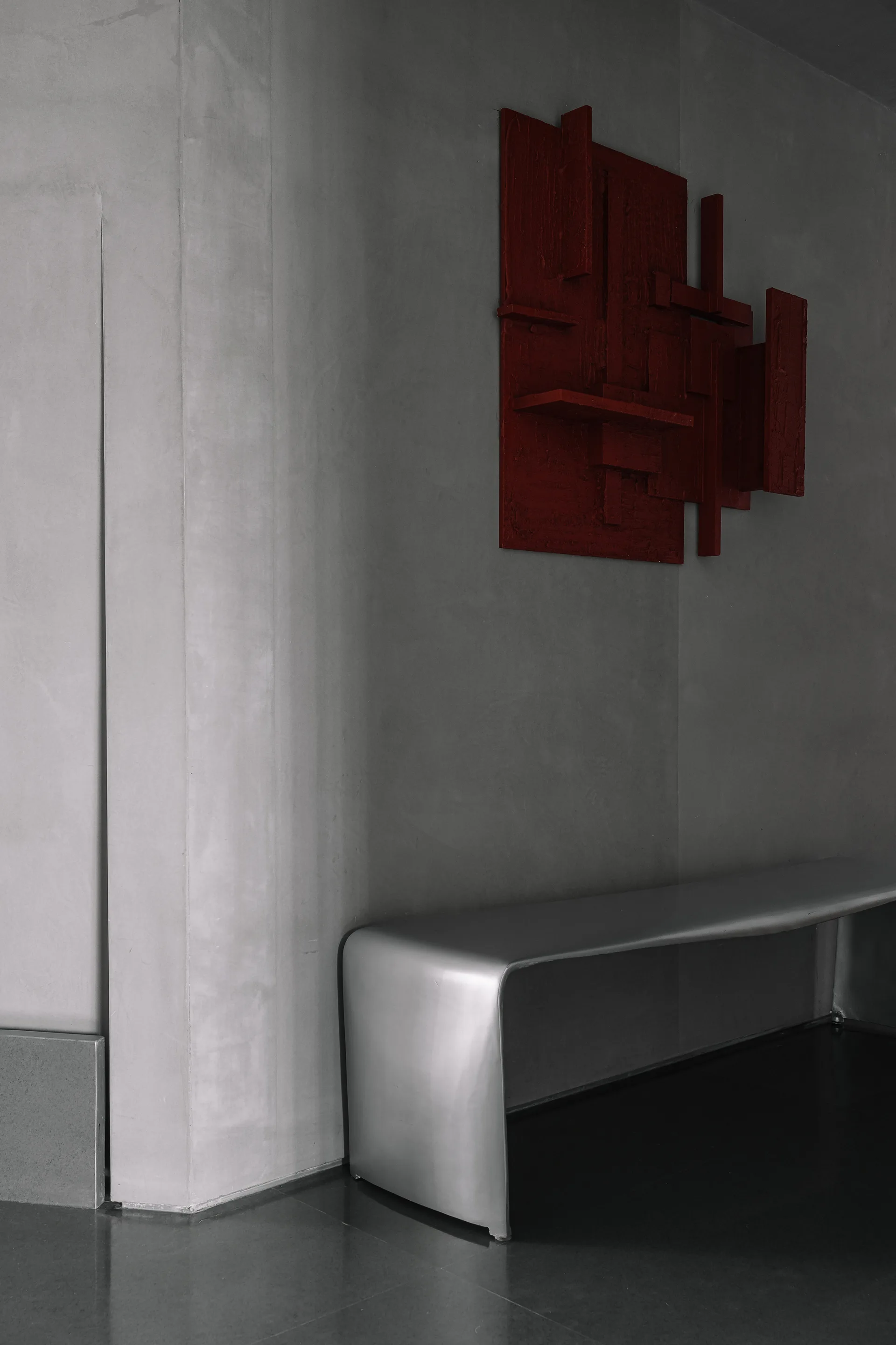 A dark red geometric wall sculpture hangs on a smooth gray plaster wall above a curved silver metal bench in a minimalist space.