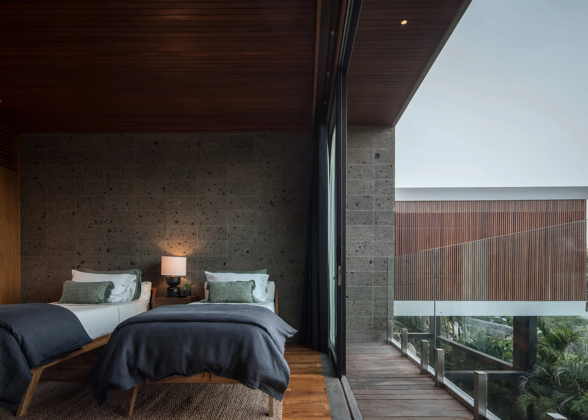 A modern bedroom with two twin beds, a textured stone wall, a wood ceiling, and sliding glass doors leading to a balcony.