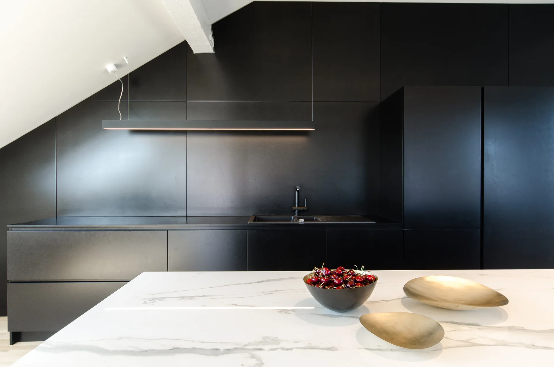 A modern kitchen featuring matte black cabinets under a sloped ceiling and a white marble-patterned island countertop with decorative brass bowls.