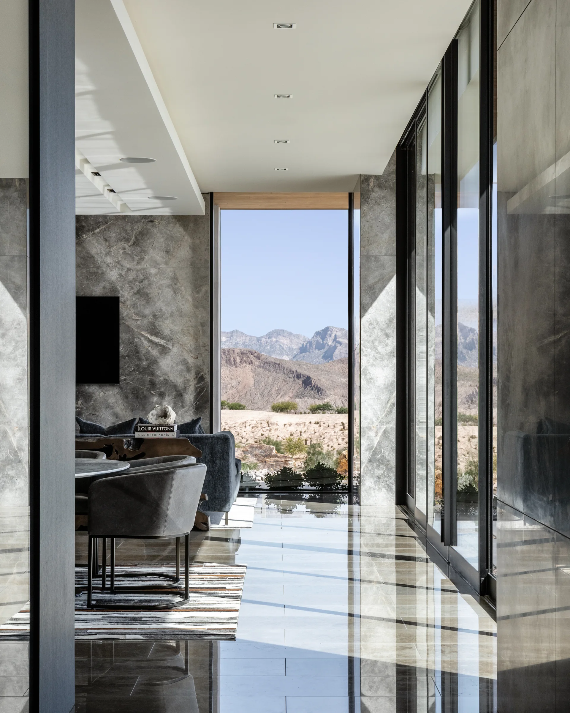 A modern living space features polished gray stone floors, veined stone walls, and floor-to-ceiling glass windows showing a desert mountain landscape.