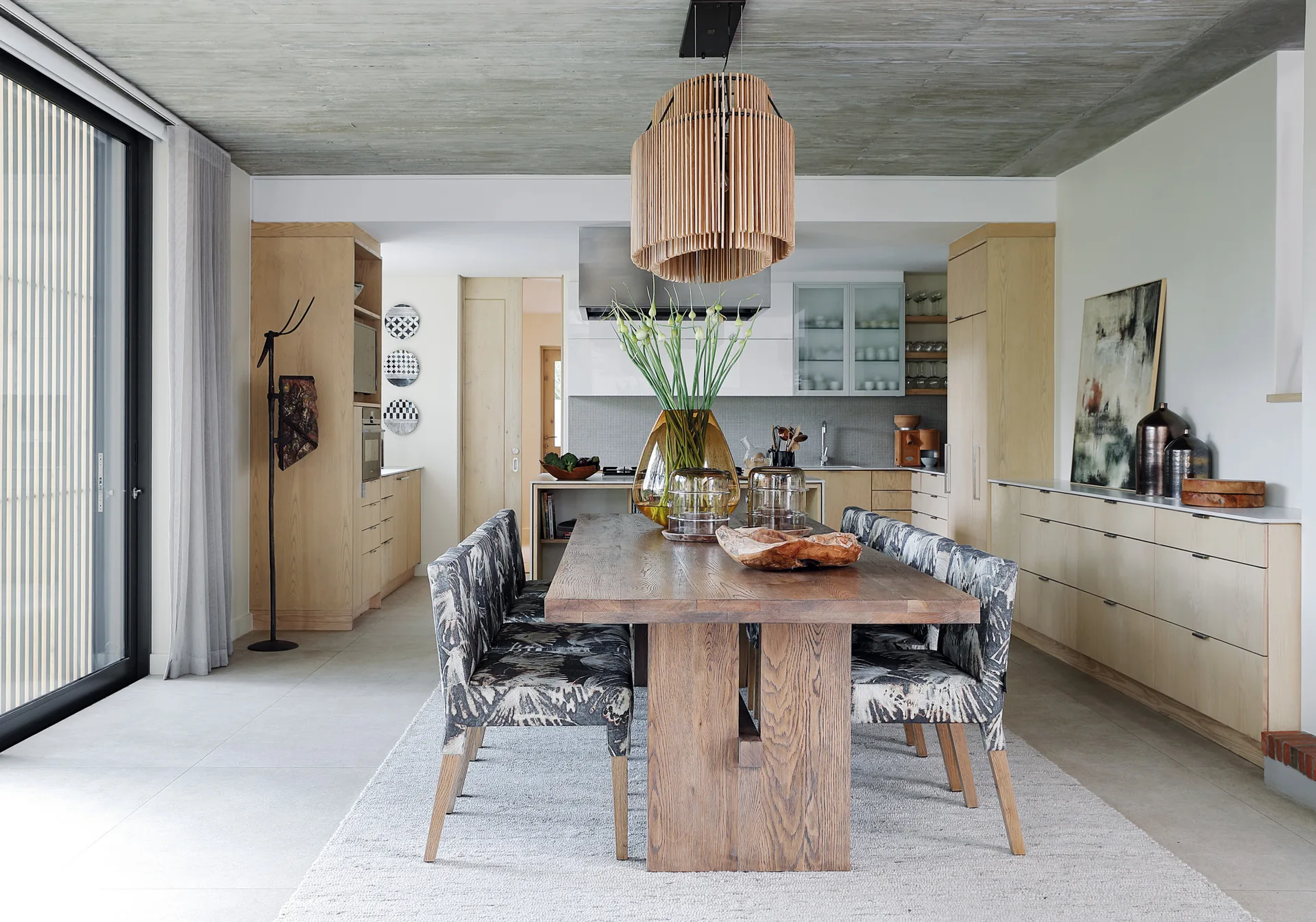 A dining room features a long wooden table surrounded by patterned fabric chairs under a large slatted wood pendant light and a concrete ceiling.