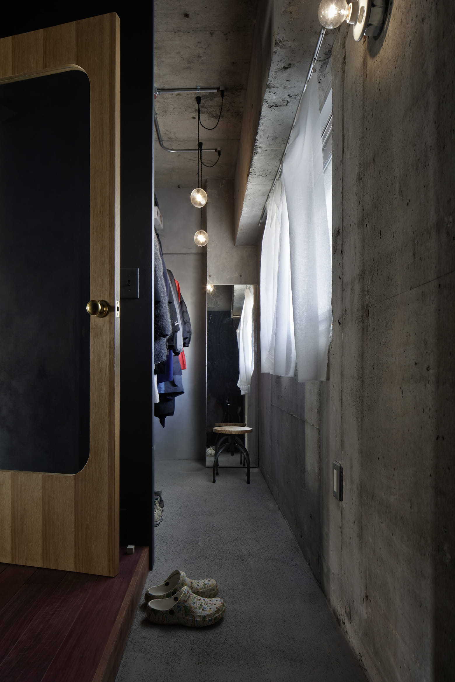 A narrow industrial walk-in closet features exposed raw concrete walls, hanging clothes on an open rack, a sheer white curtain, and pendant lighting.