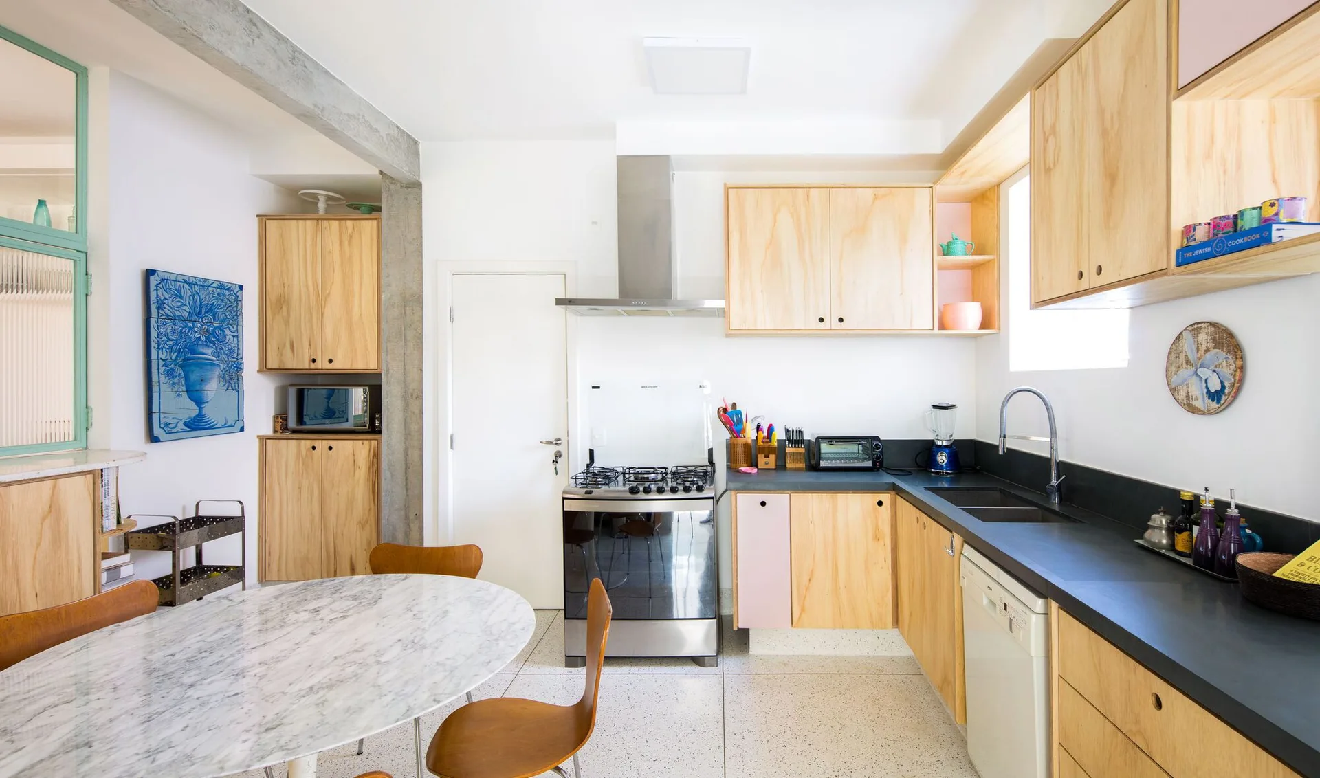 A bright kitchen features light wood plywood cabinets, black countertops, an exposed concrete ceiling beam, and a marble table.