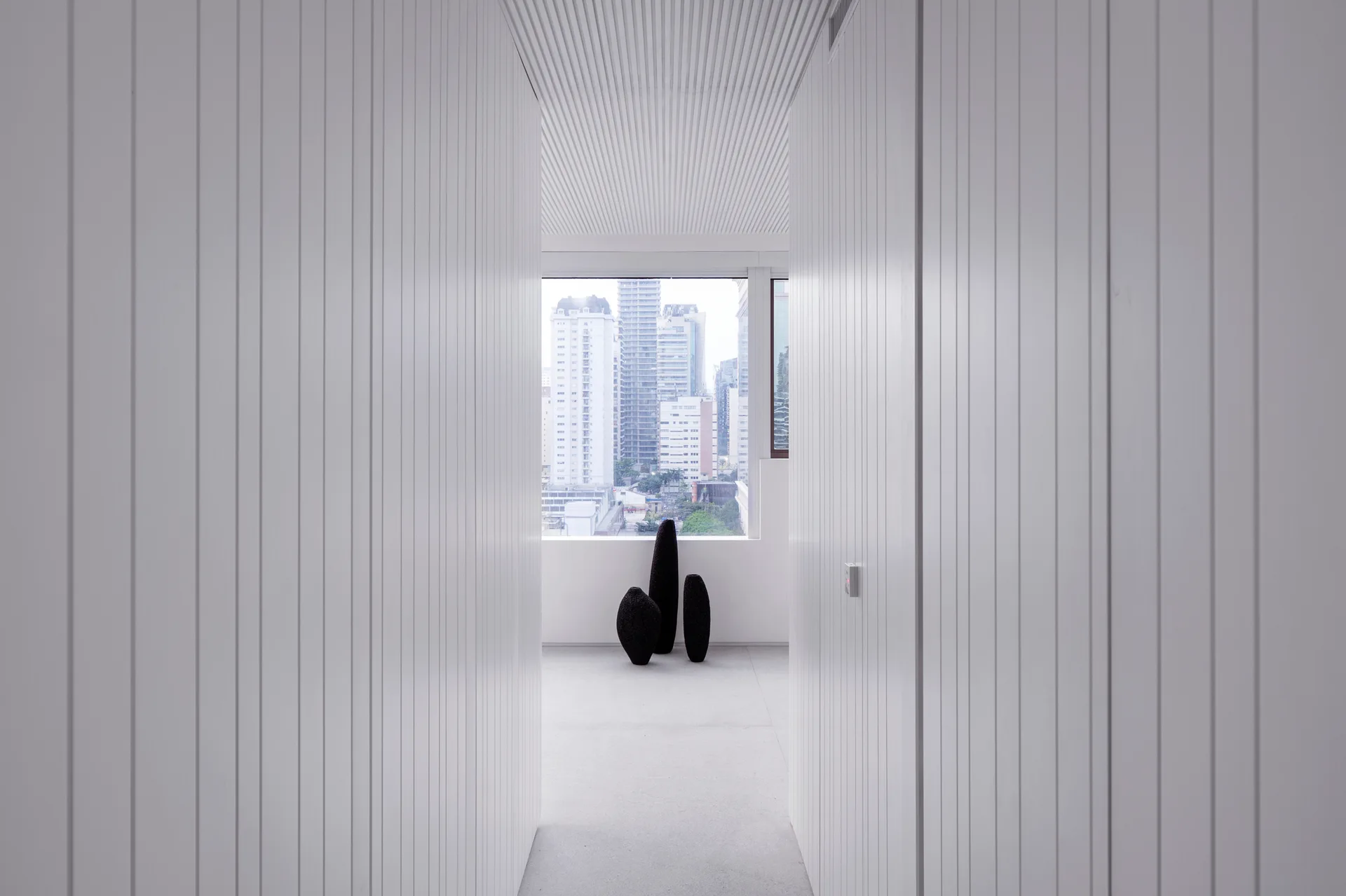 A narrow white hallway with vertical wall slats and a slatted ceiling leads to a window overlooking city buildings, featuring three dark floor sculptures.