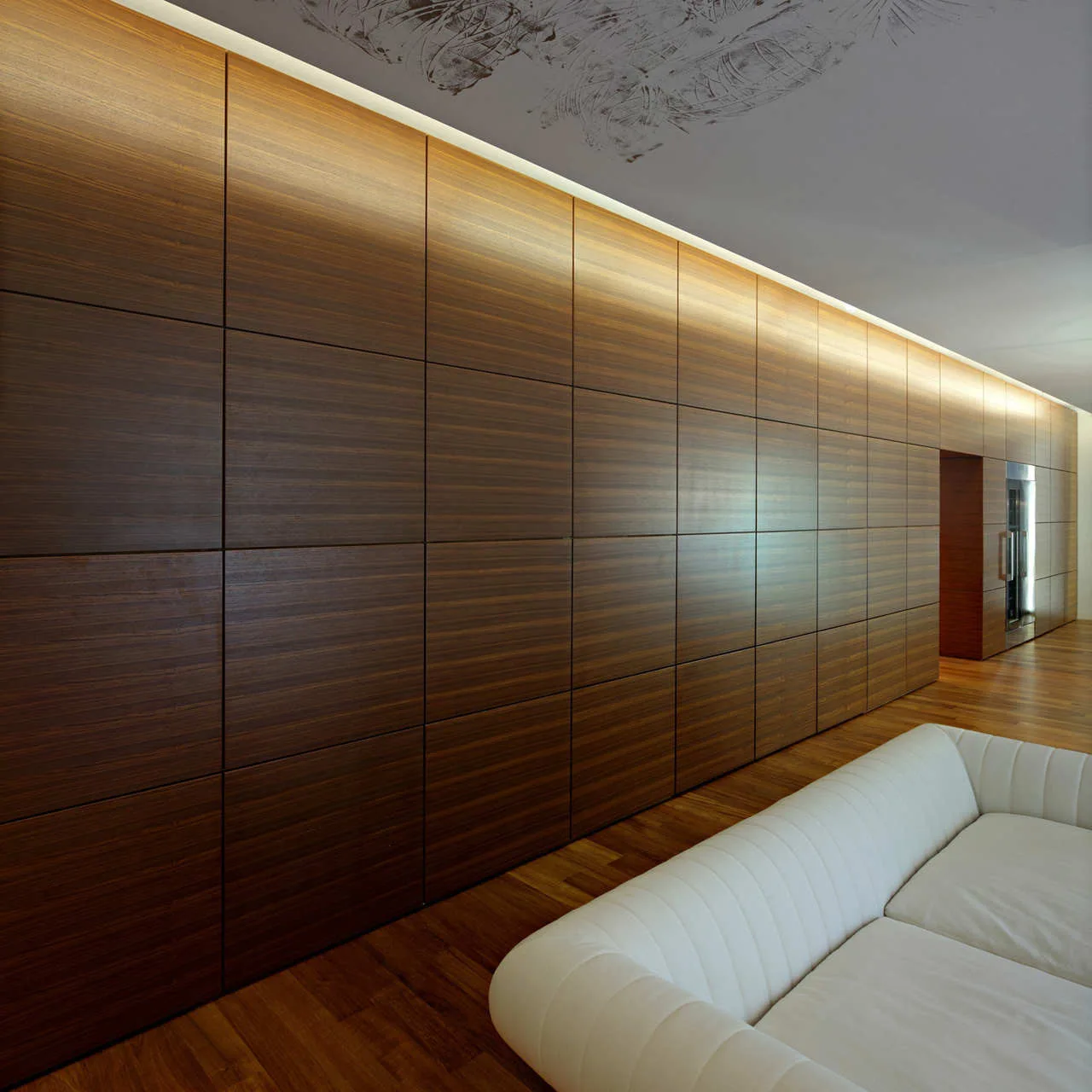 A modern living room features a full-length dark wood paneled wall lit by overhead cove lighting next to a white channeled sofa on wood floors.
