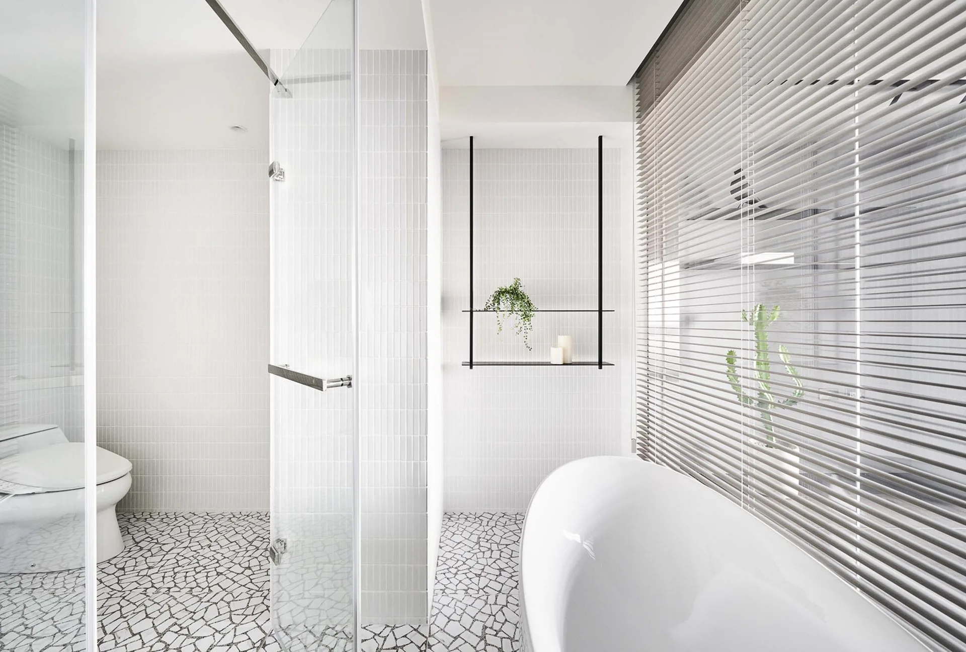 A minimalist white bathroom featuring a freestanding tub, glass shower screen, vertical subway tile walls, and broken-stone mosaic tile flooring.