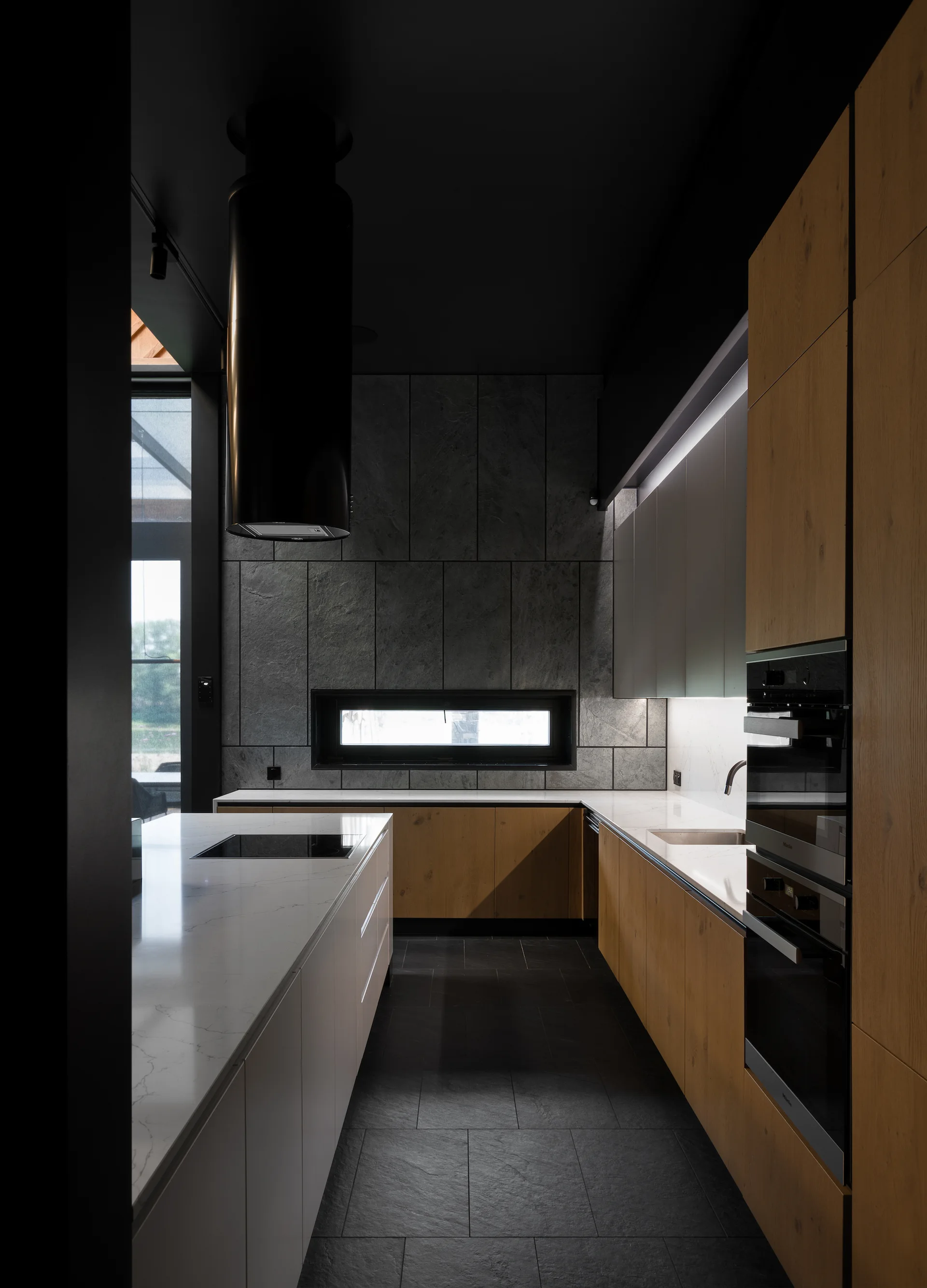 A modern kitchen features wood cabinets, white countertops, a dark tiled floor, a stone block accent wall, and a black cylindrical range hood.