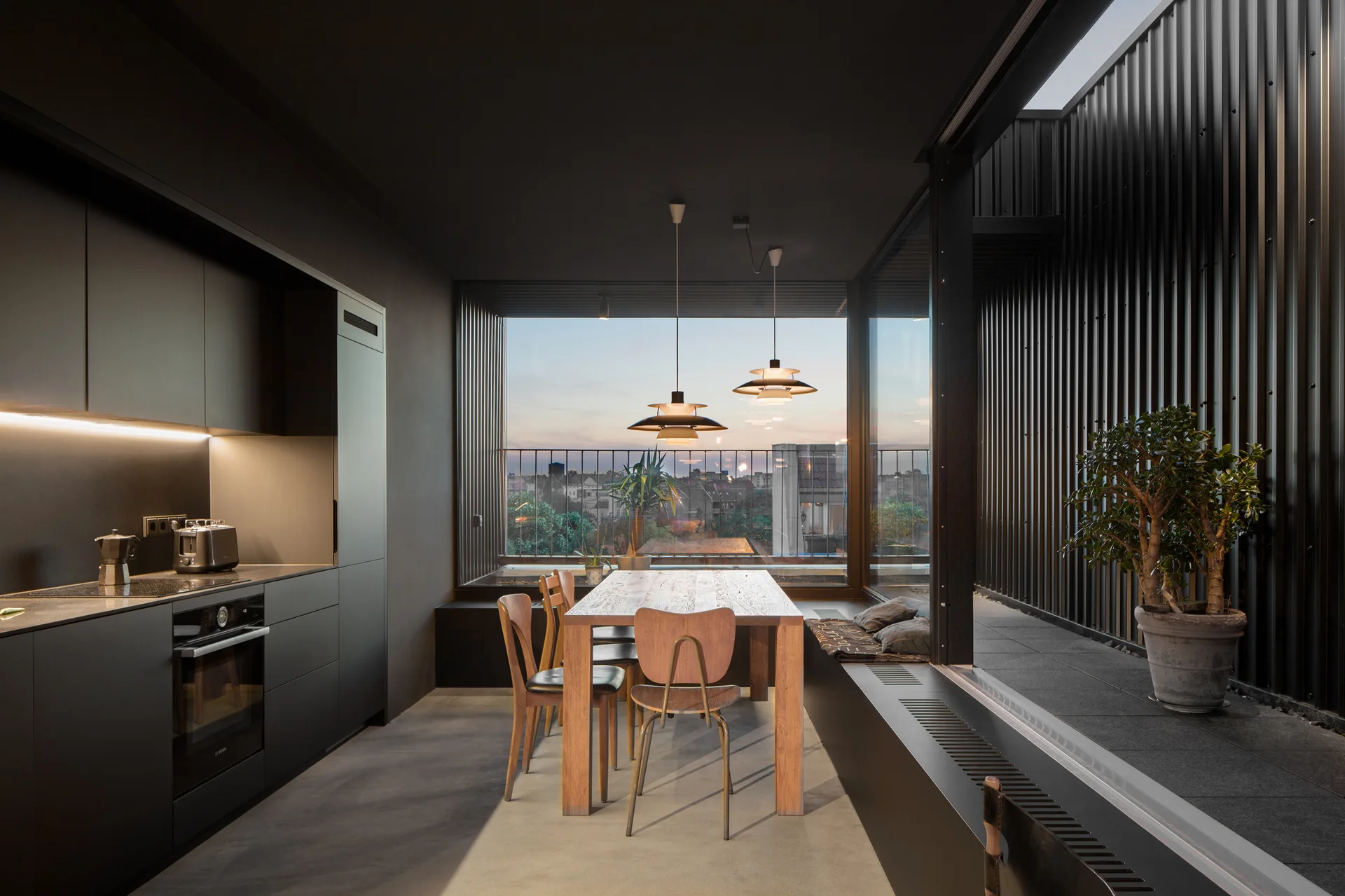 A modern kitchen with matte black cabinets, a wooden dining table, and large glass windows opening to a dark metal balcony at dusk.