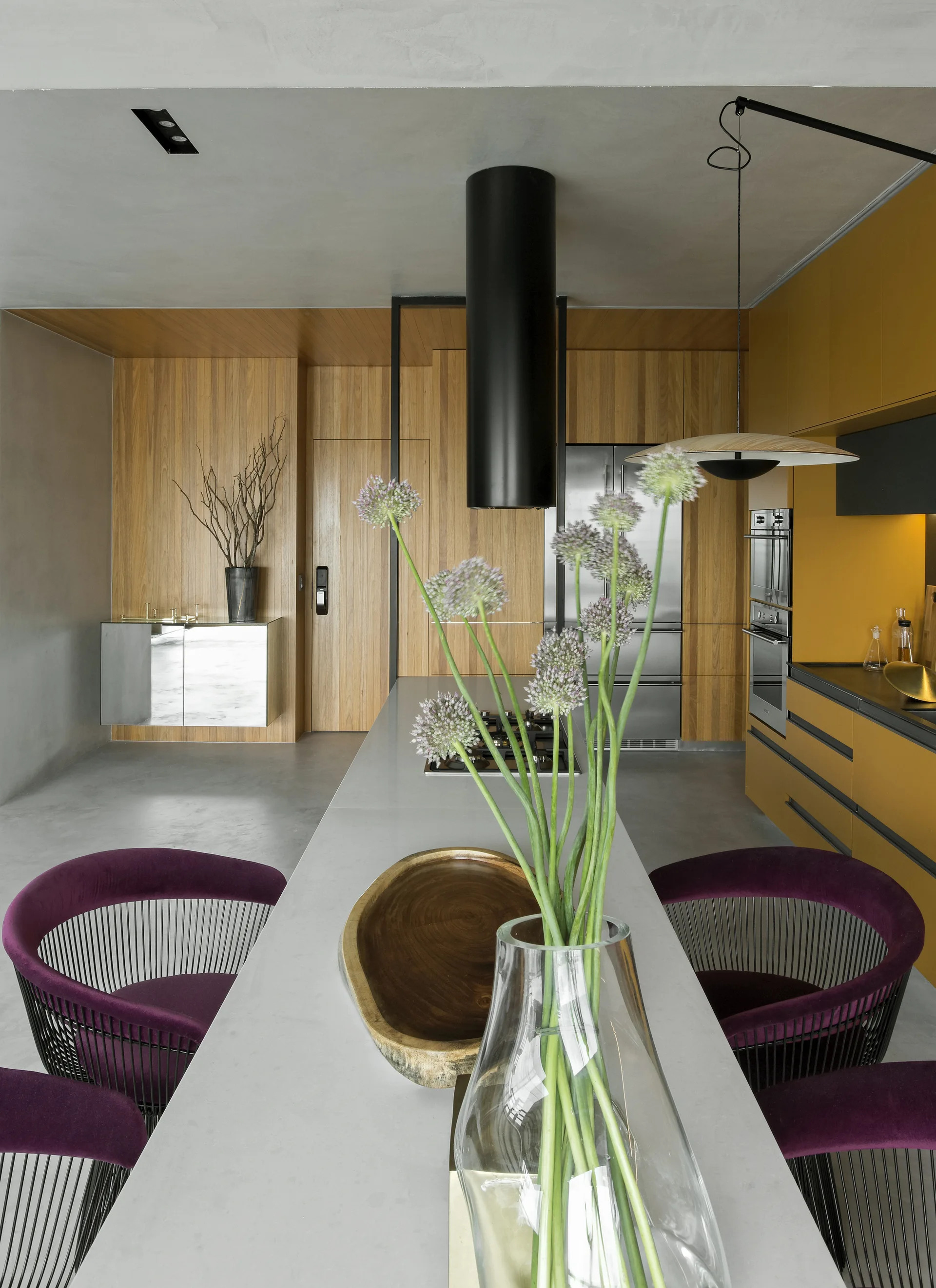 A modern residential kitchen features a grey concrete island with purple chairs, mustard yellow cabinets, and warm wood wall paneling.