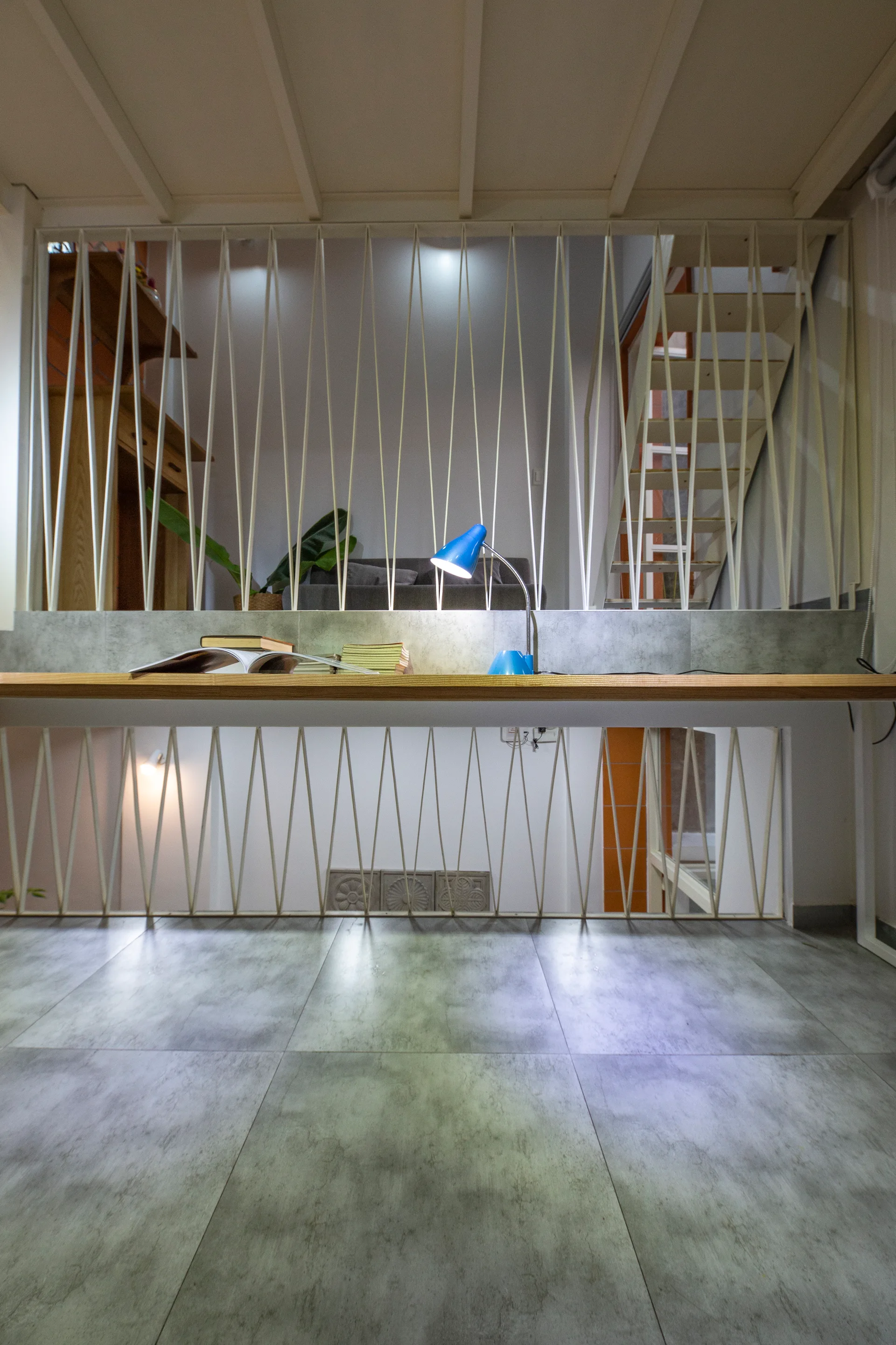 A wooden desk with a blue lamp sits in front of a white geometric metal screen partitioning a multi-level modern interior with grey tiled floors.