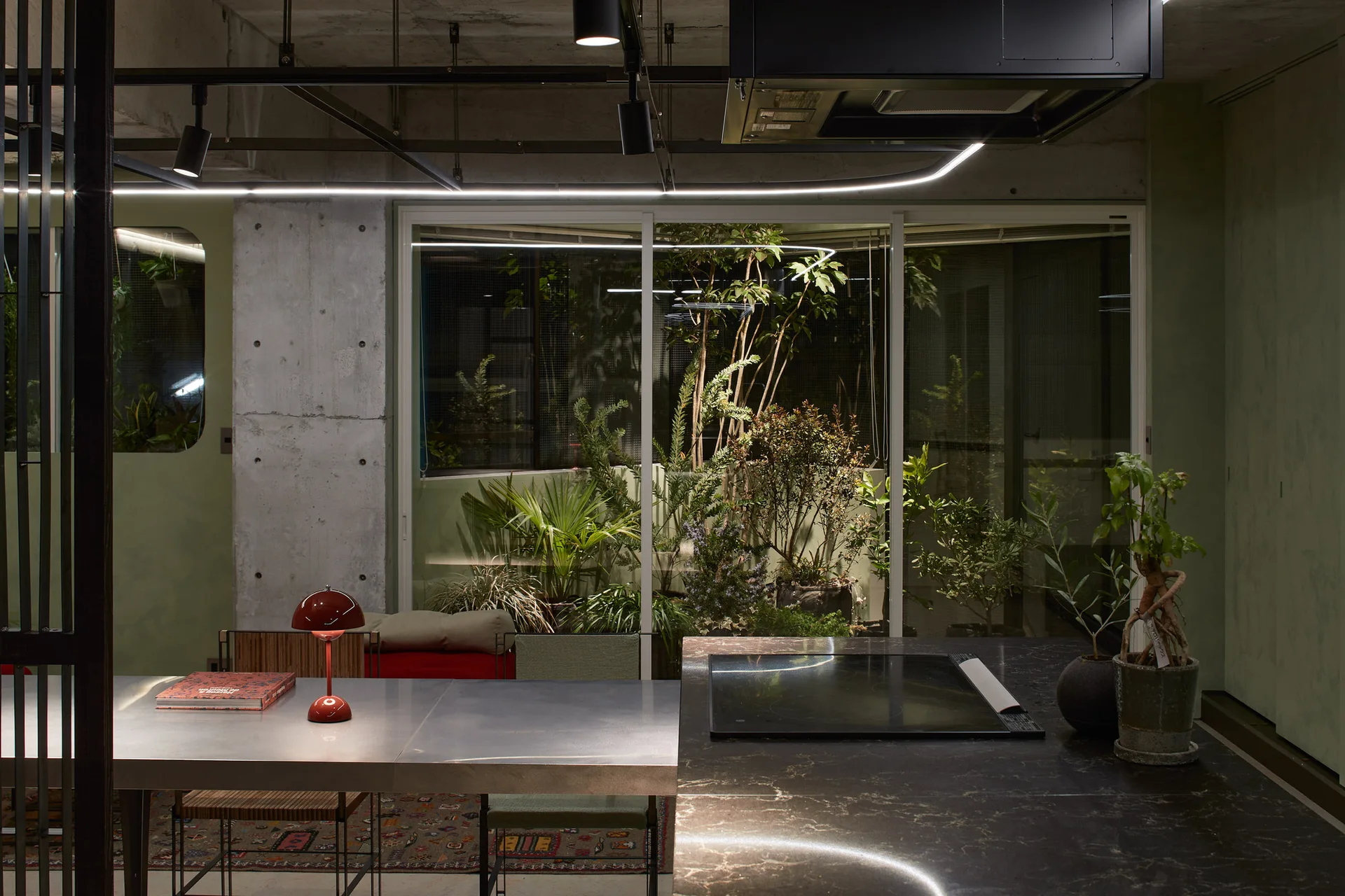 A modern kitchen with a dark veined stone island and a metallic dining table next to a large glass window overlooking a plant-filled courtyard.