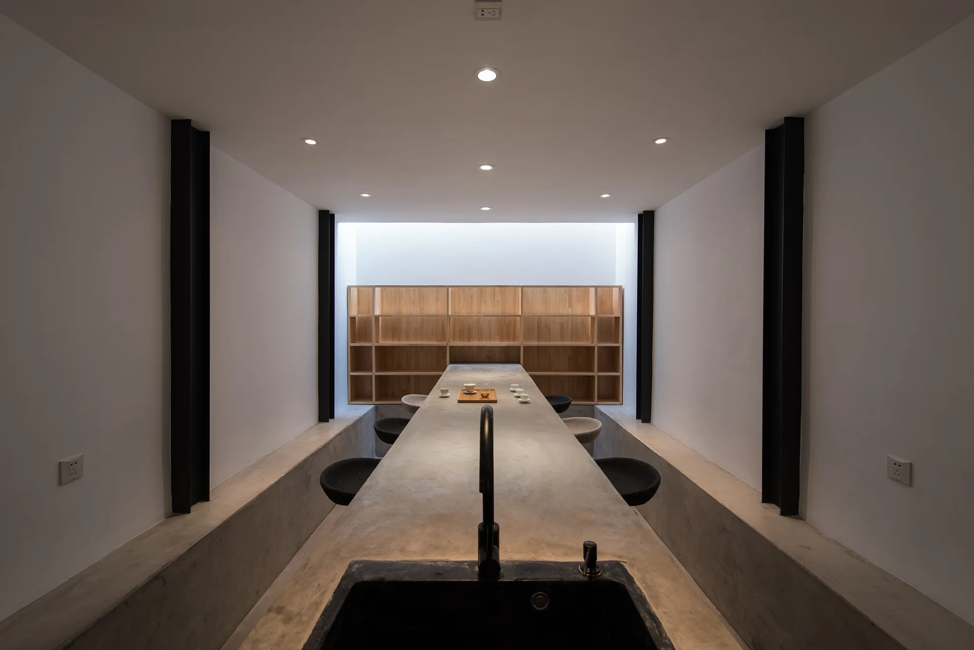 A long concrete communal table with a sink in a symmetrical minimalist room flanked by sunken floor seating and a wooden grid bookshelf.