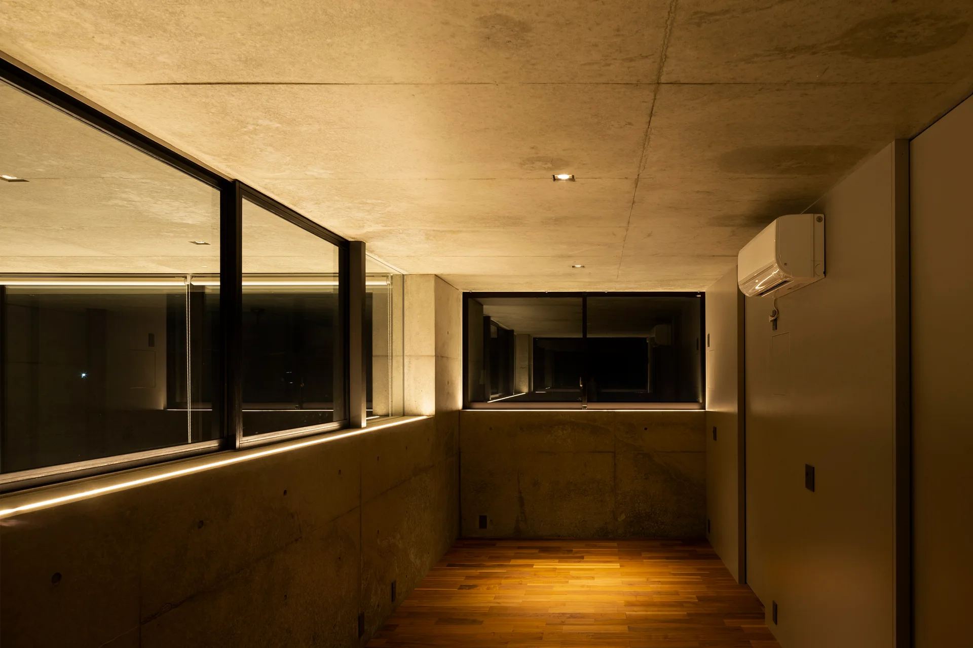 A modern concrete corridor features warm wood plank flooring, black-framed windows, and built-in linear accent lighting glowing beneath the sills.
