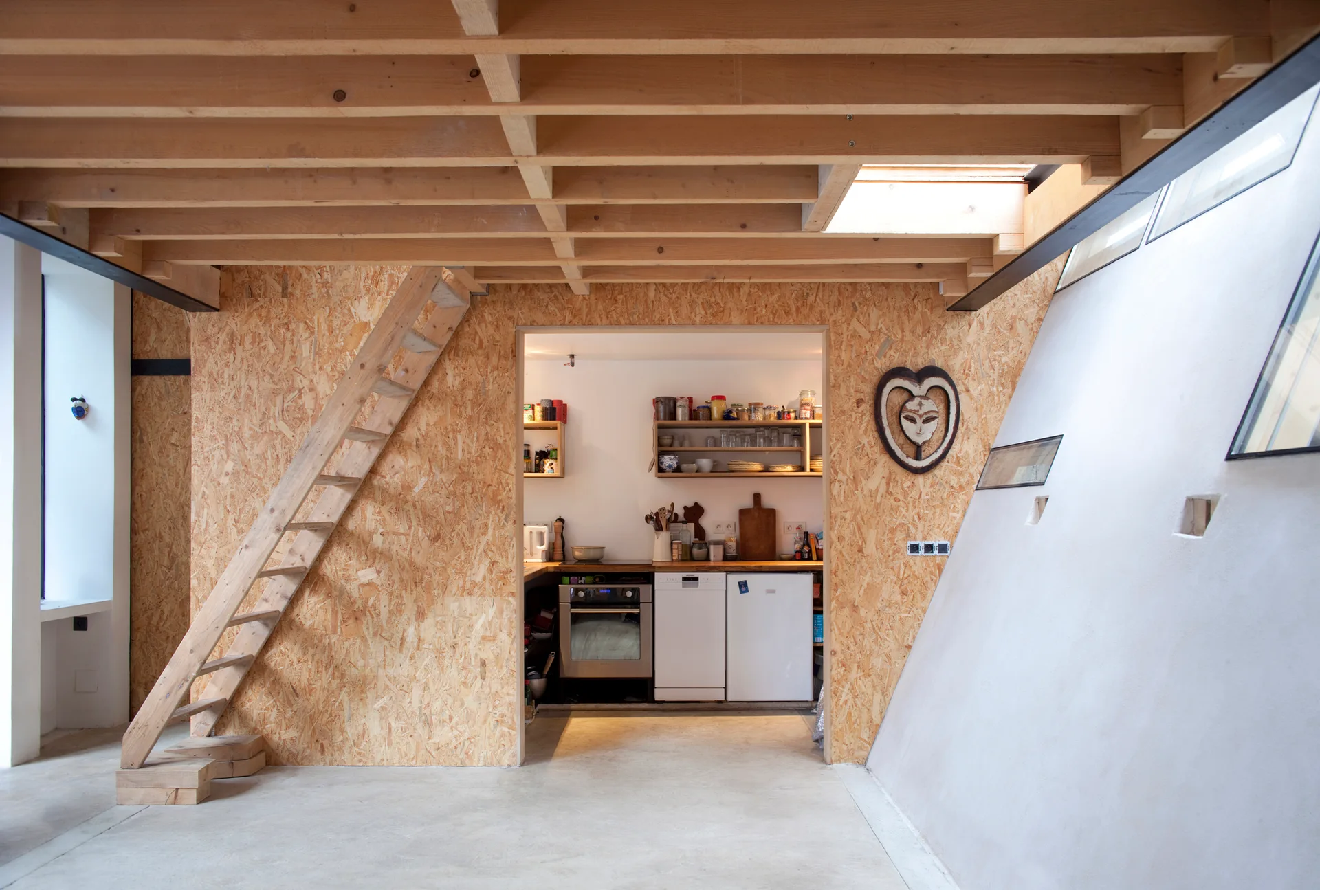A minimalist loft space features walls made of raw OSB panels, an exposed timber beam ceiling, a wooden ladder, and a kitchen visible through a cutout.