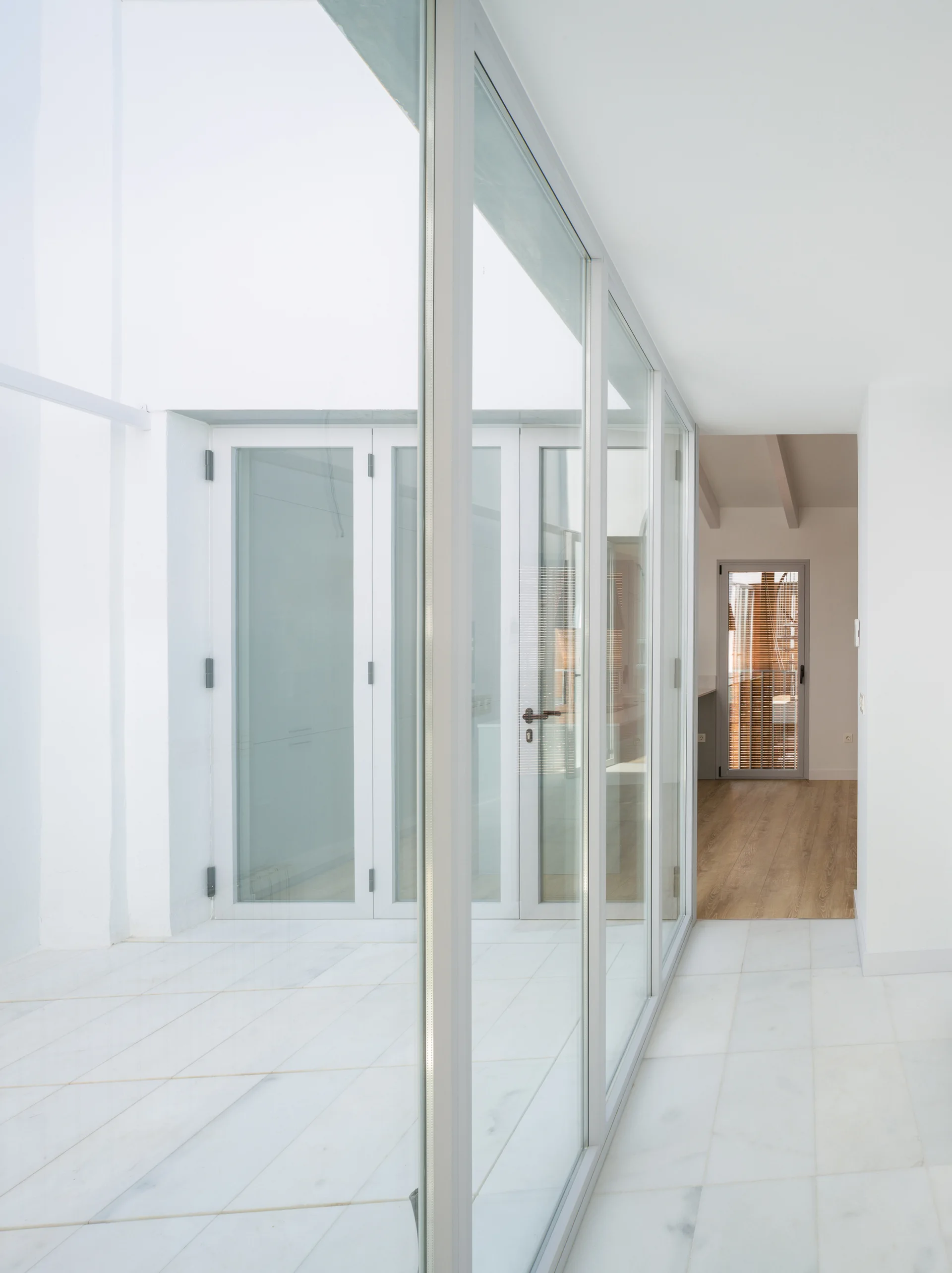 A bright minimalist hallway featuring white marble tile flooring and a large glass partition wall with slim metal frames.