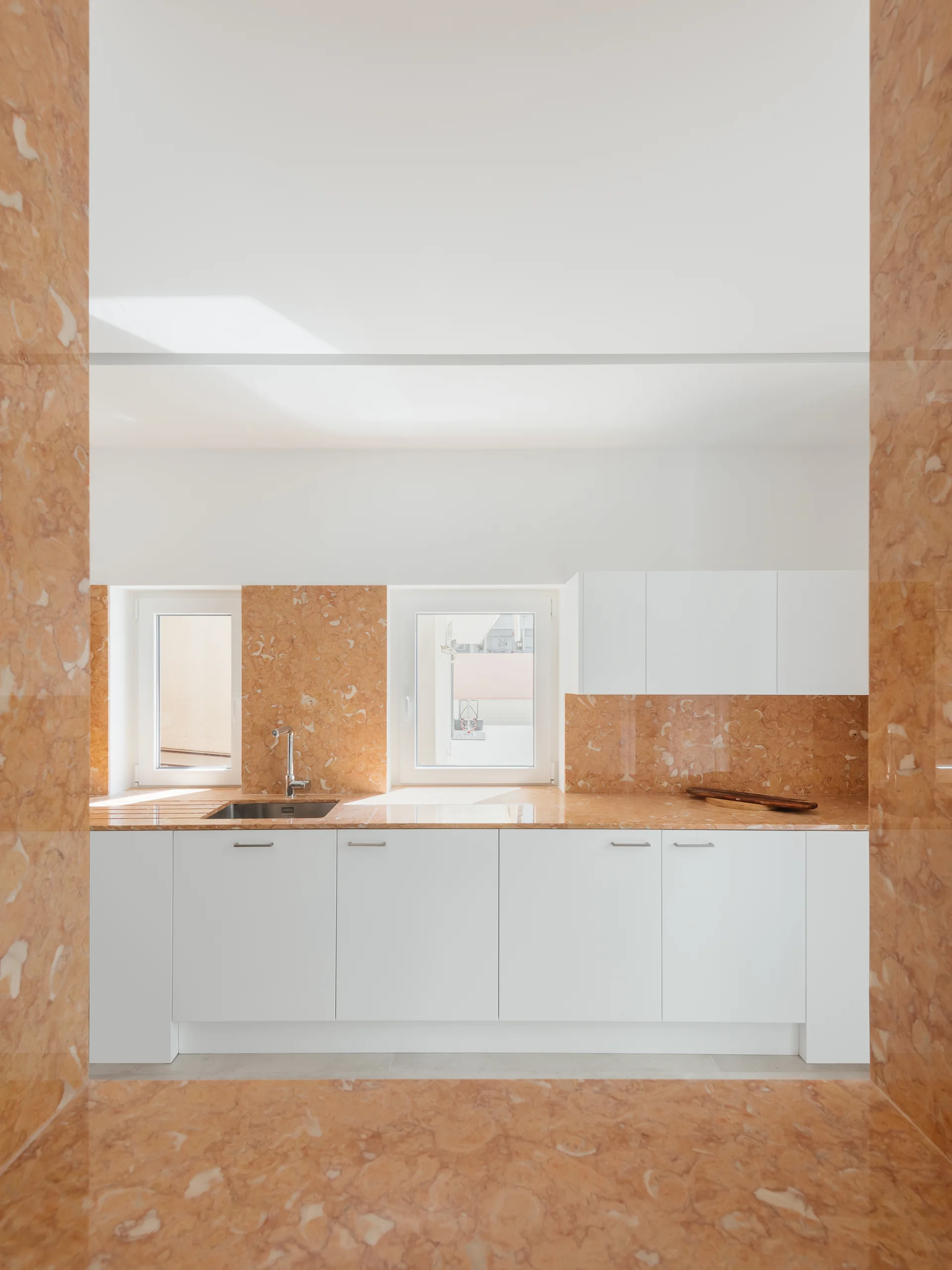A minimalist kitchen featuring white lower cabinetry, a stainless steel sink, and polished orange marble countertops and wall cladding.