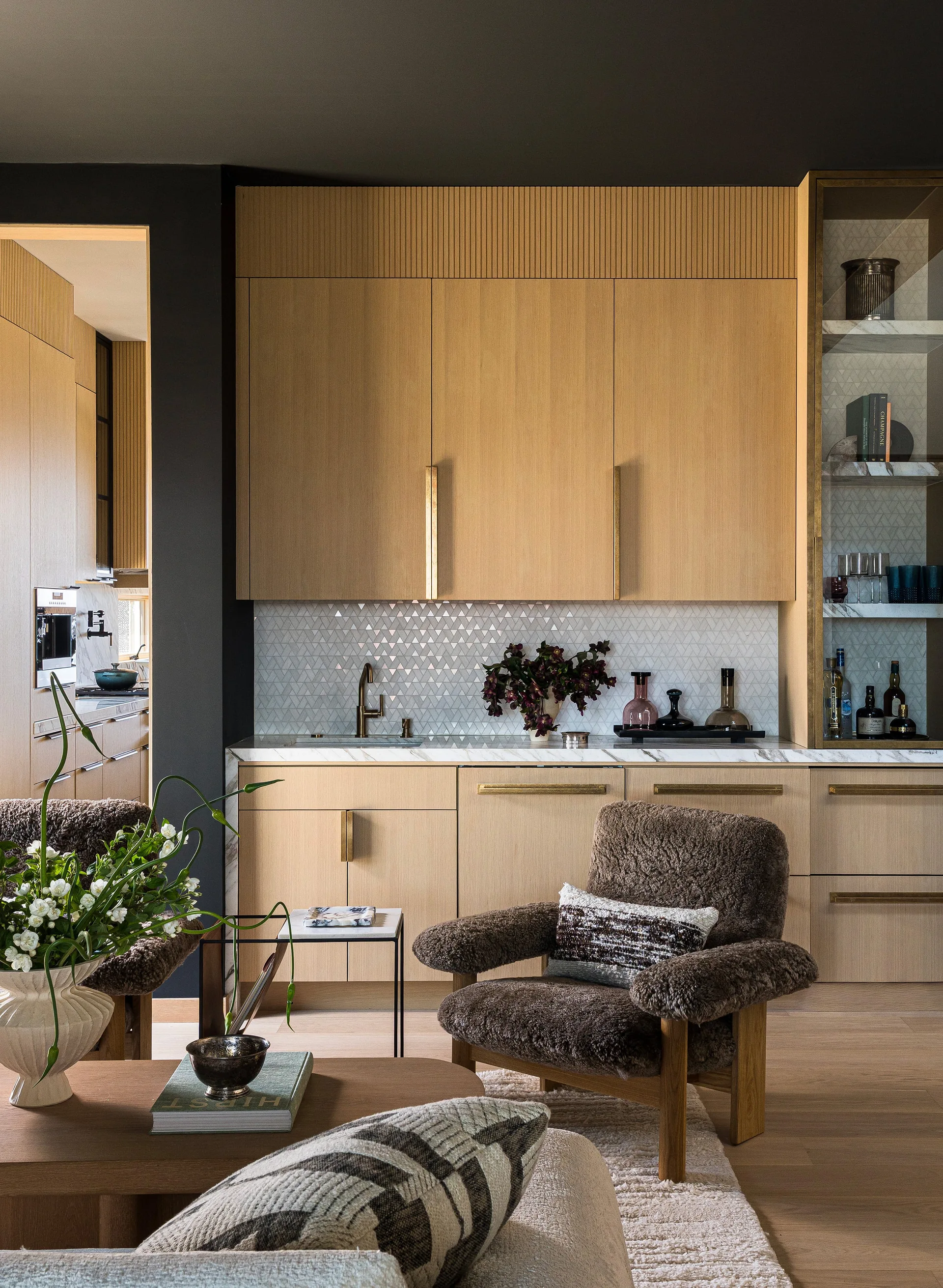A home wet bar with light wood cabinets, a geometric mosaic tile backsplash, and a marble countertop sits behind a fuzzy brown armchair.