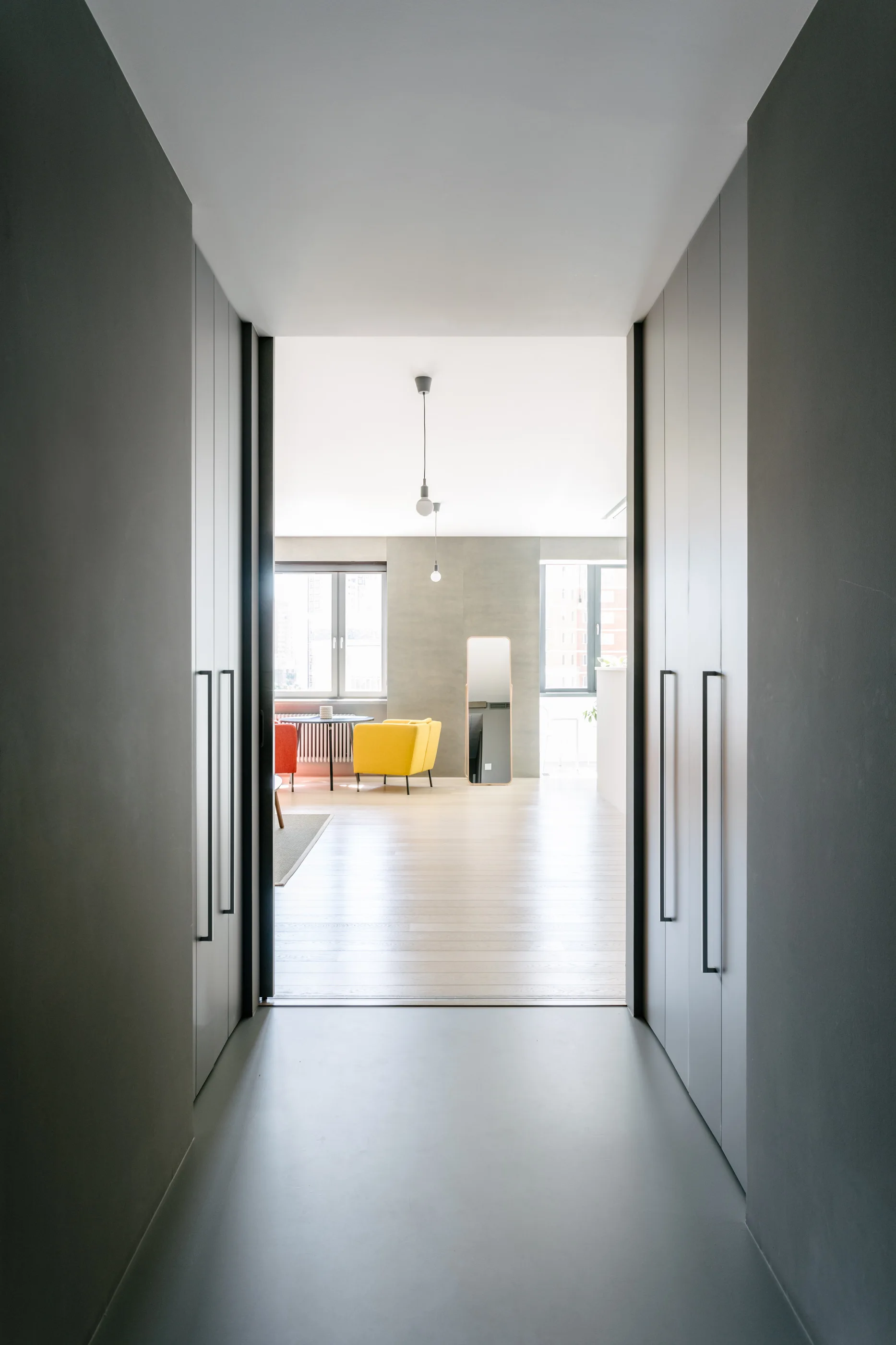 A view through a gray hallway lined with built-in cabinets looking into a bright living room with a yellow armchair and wood flooring.