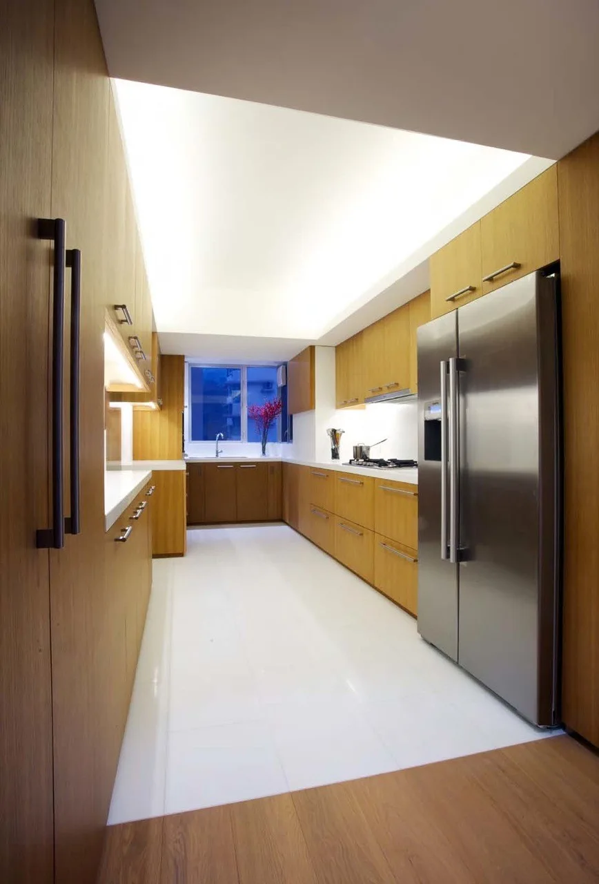 A modern galley kitchen features flat-front wood cabinetry, a stainless steel refrigerator, white floors, and a bright illuminated ceiling panel.
