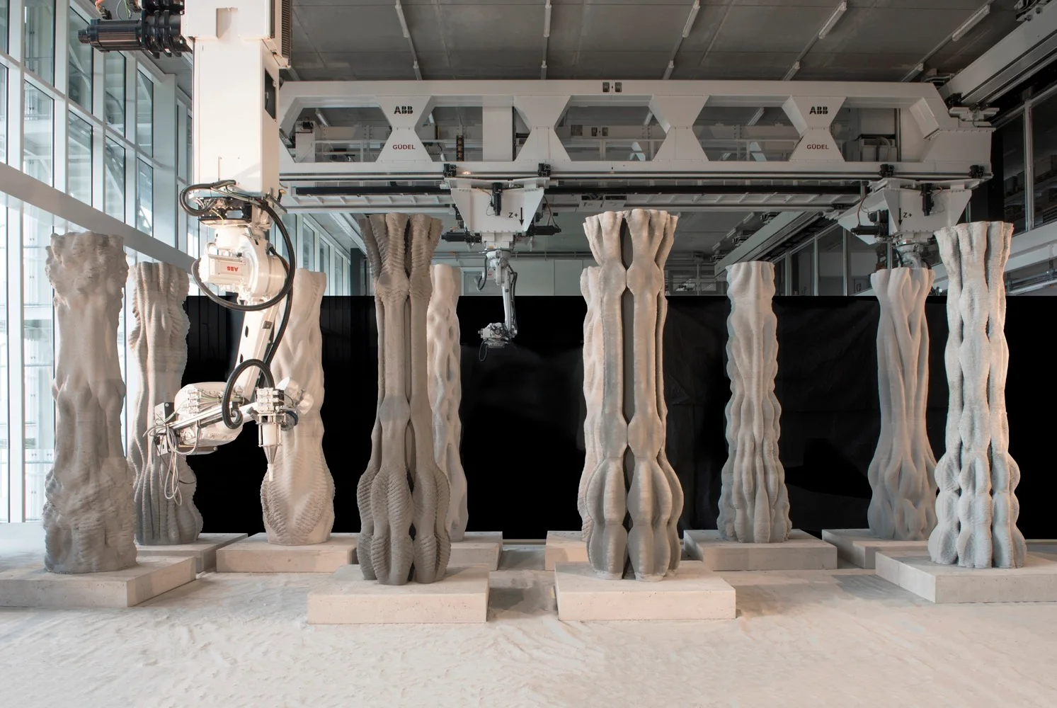 Several large grey 3D-printed concrete columns with textured organic shapes stand in an industrial facility near a robotic arm.