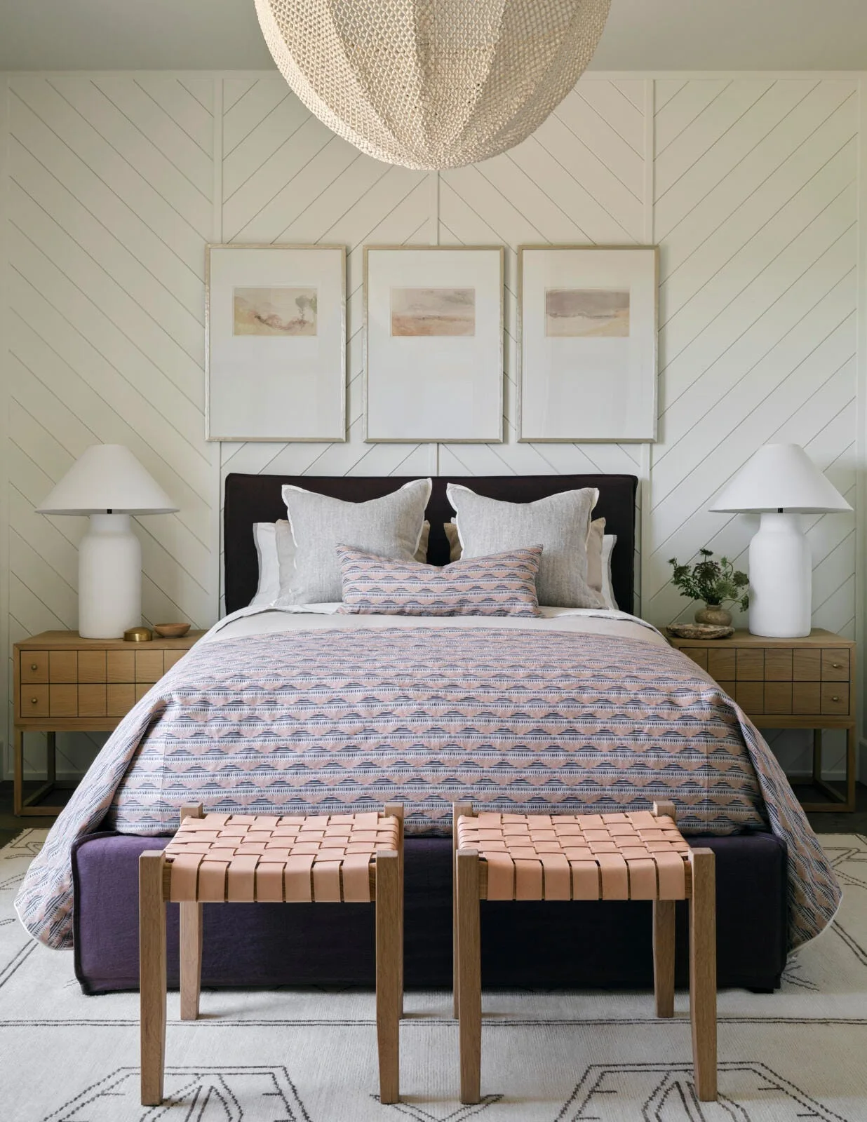 A centered double bed with a patterned coverlet sits in front of a white wall with diagonal paneling, flanked by wood nightstands and white lamps.