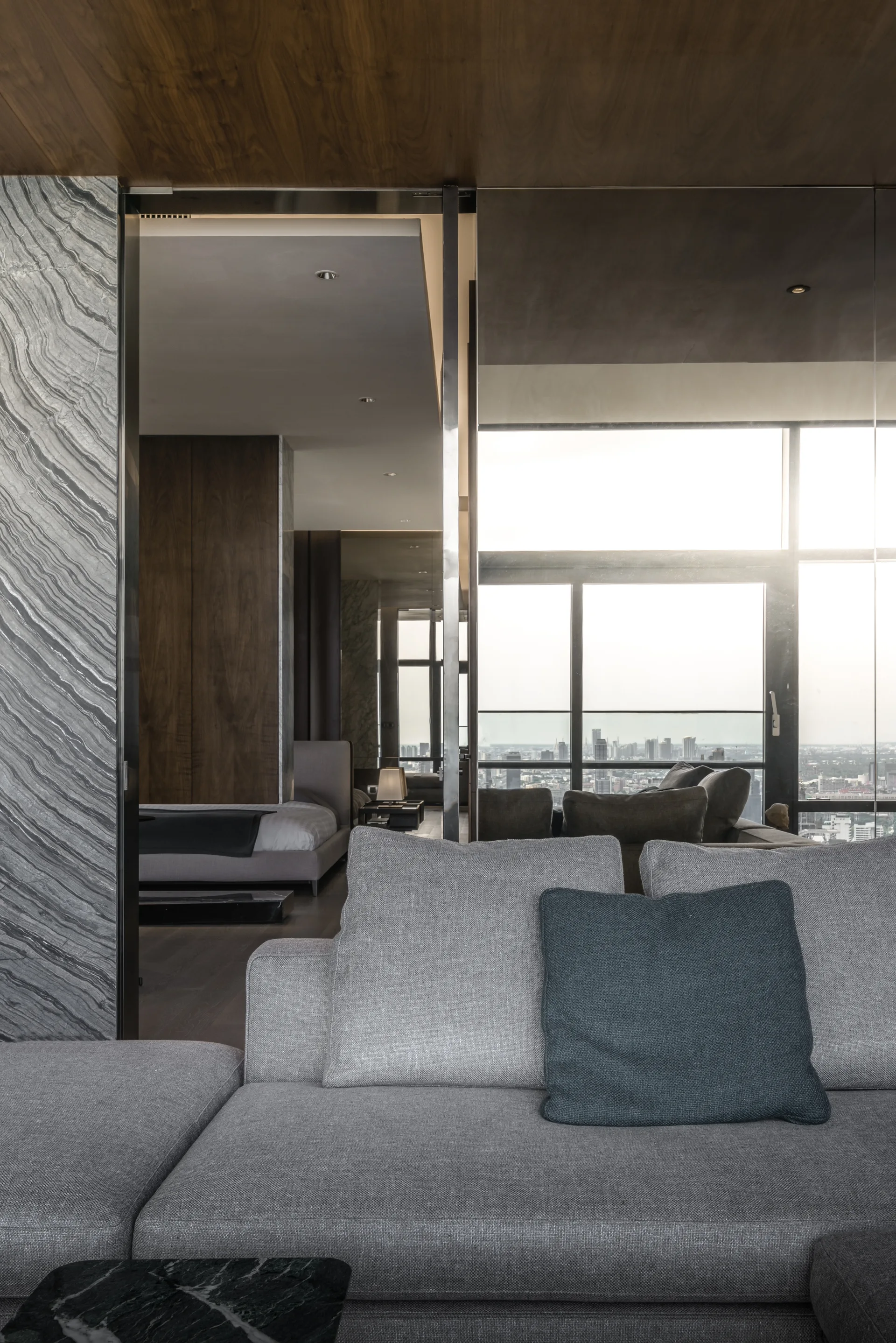 A modern grey sectional sofa sits near a veined stone wall, looking through a doorway toward a bedroom and floor-to-ceiling city views.