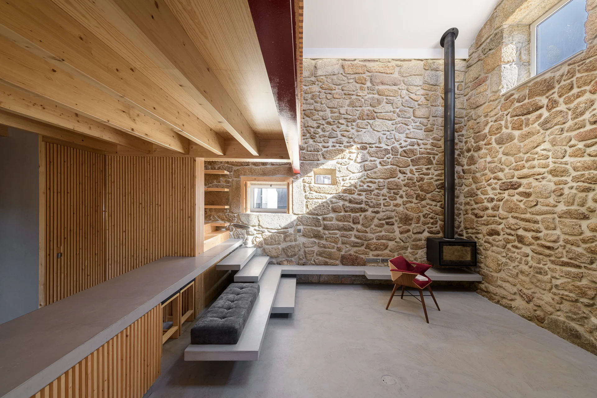 A minimalist living space featuring a high stone masonry wall with a fireplace, concrete flooring, and built-in wooden ceiling structures.