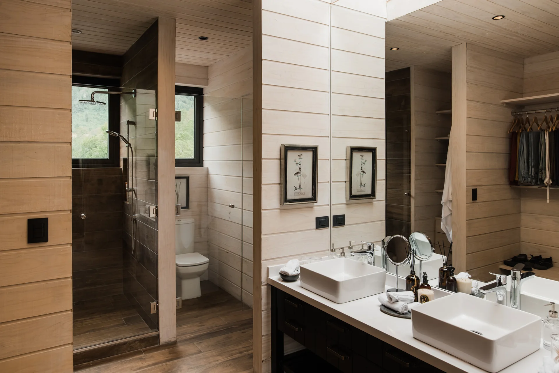 A modern rustic bathroom features horizontal light wood wall paneling, a dark vanity with two white vessel sinks, and a glass-enclosed shower.