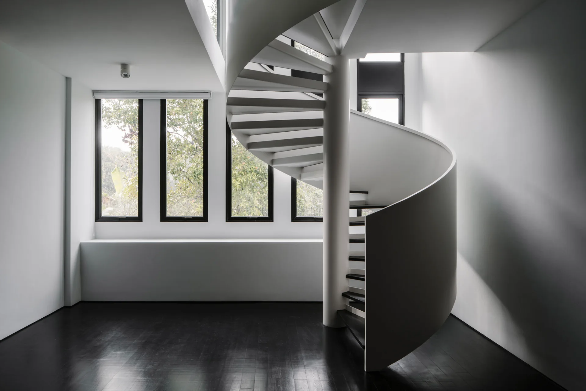 A minimalist white room features a white metal spiral staircase situated on dark wood floors next to a row of four black-framed windows.