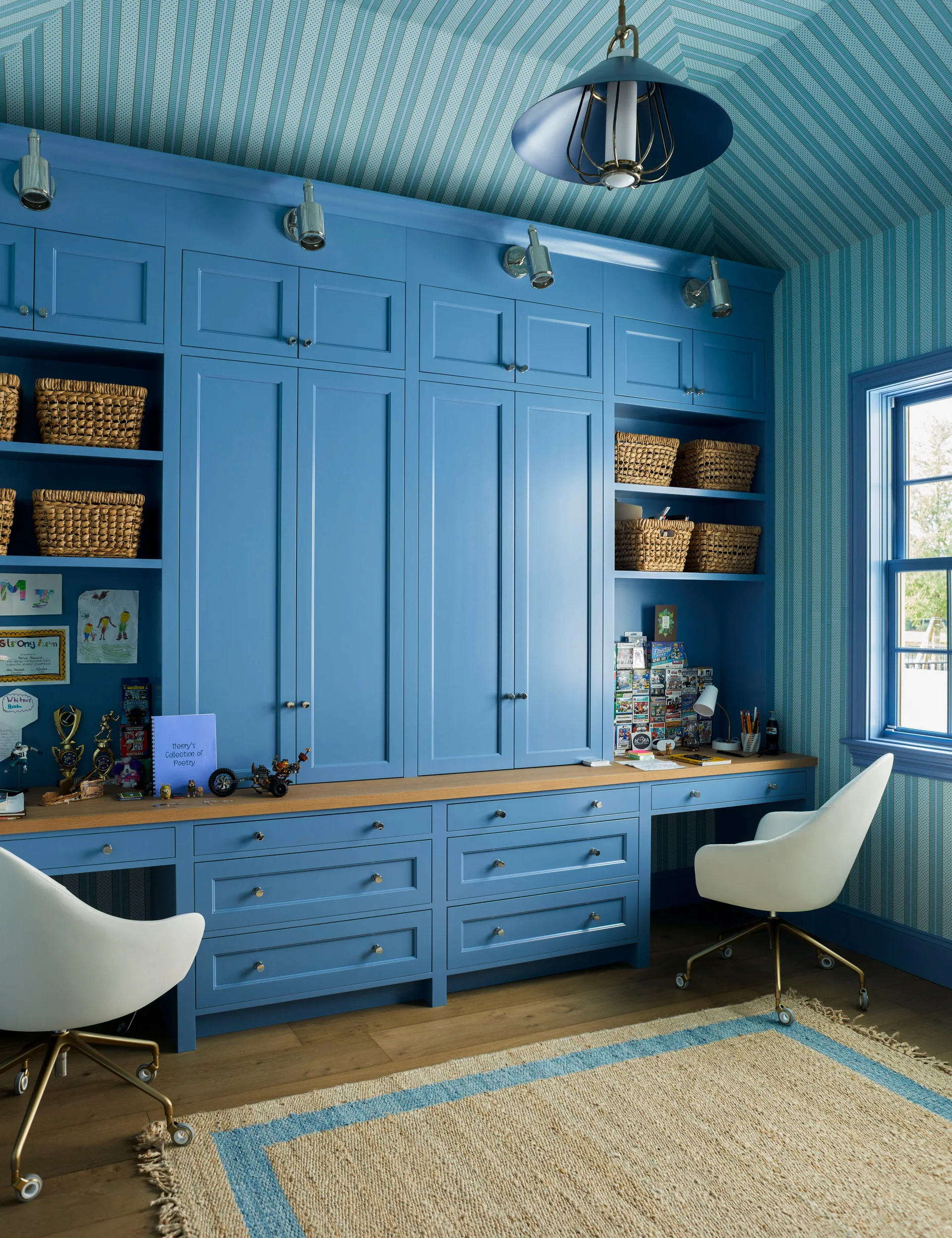 A home office featuring blue built-in cabinets with two desks, white swivel chairs, and vertical striped wallpaper covering the walls and vaulted ceiling.