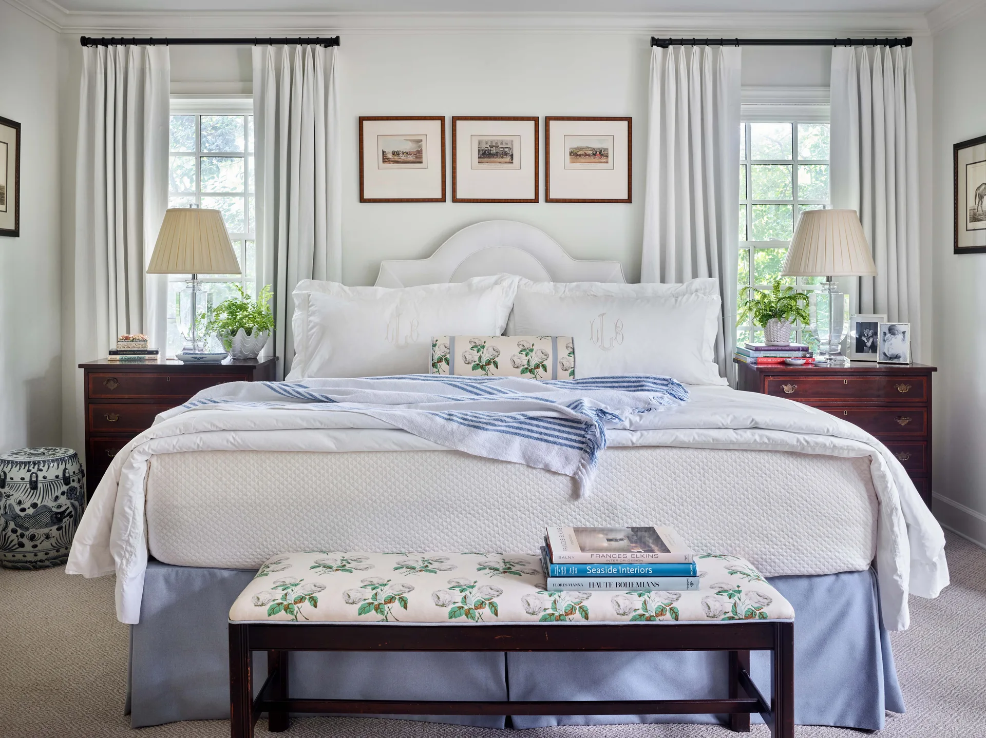 A symmetrical traditional bedroom features an upholstered white bed flanked by dark wood nightstands and windows with white curtains.