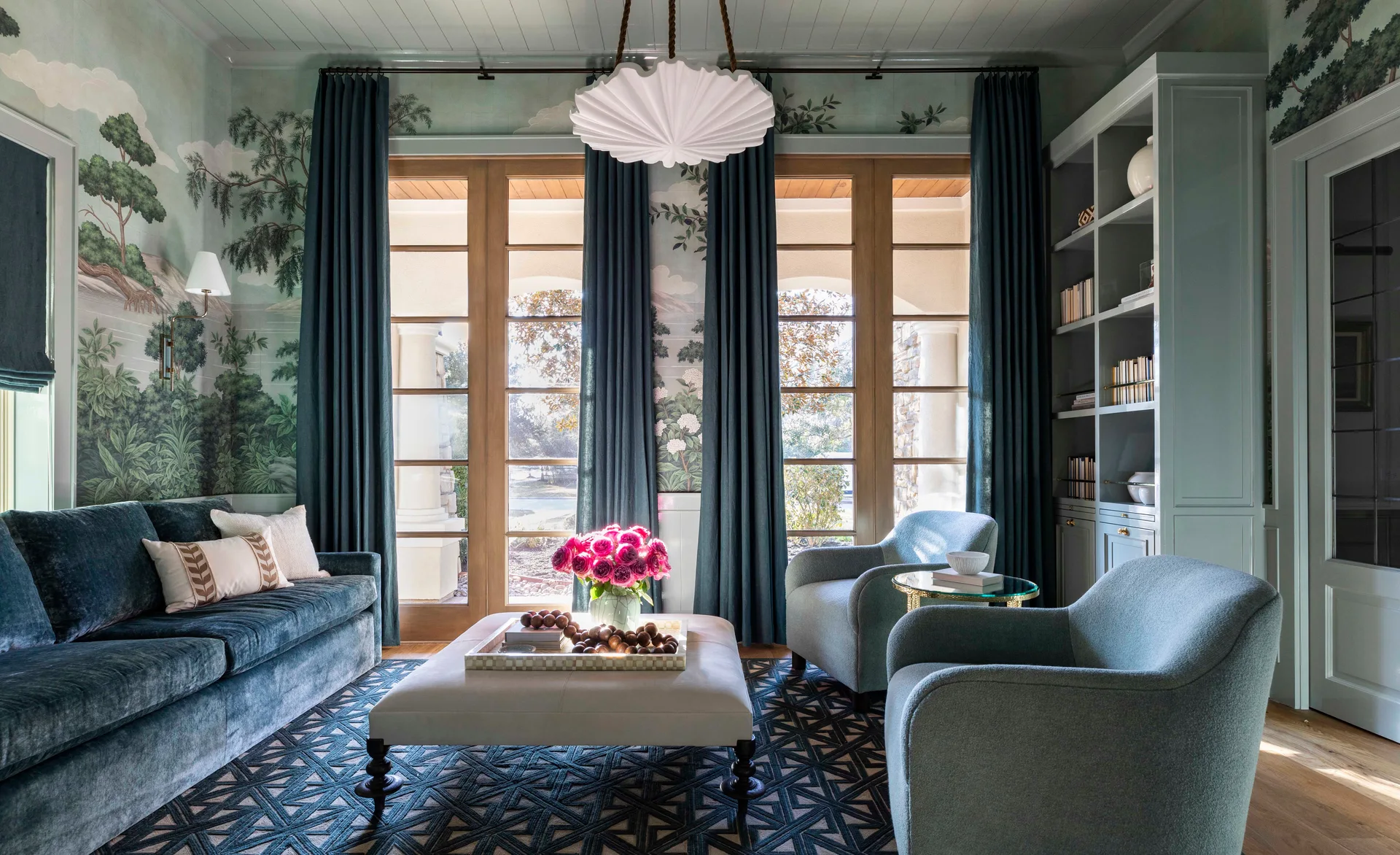 A teal living room with a scenic mural on the walls, full-length drapes, a velvet sofa, and a scalloped pendant light over an ottoman.