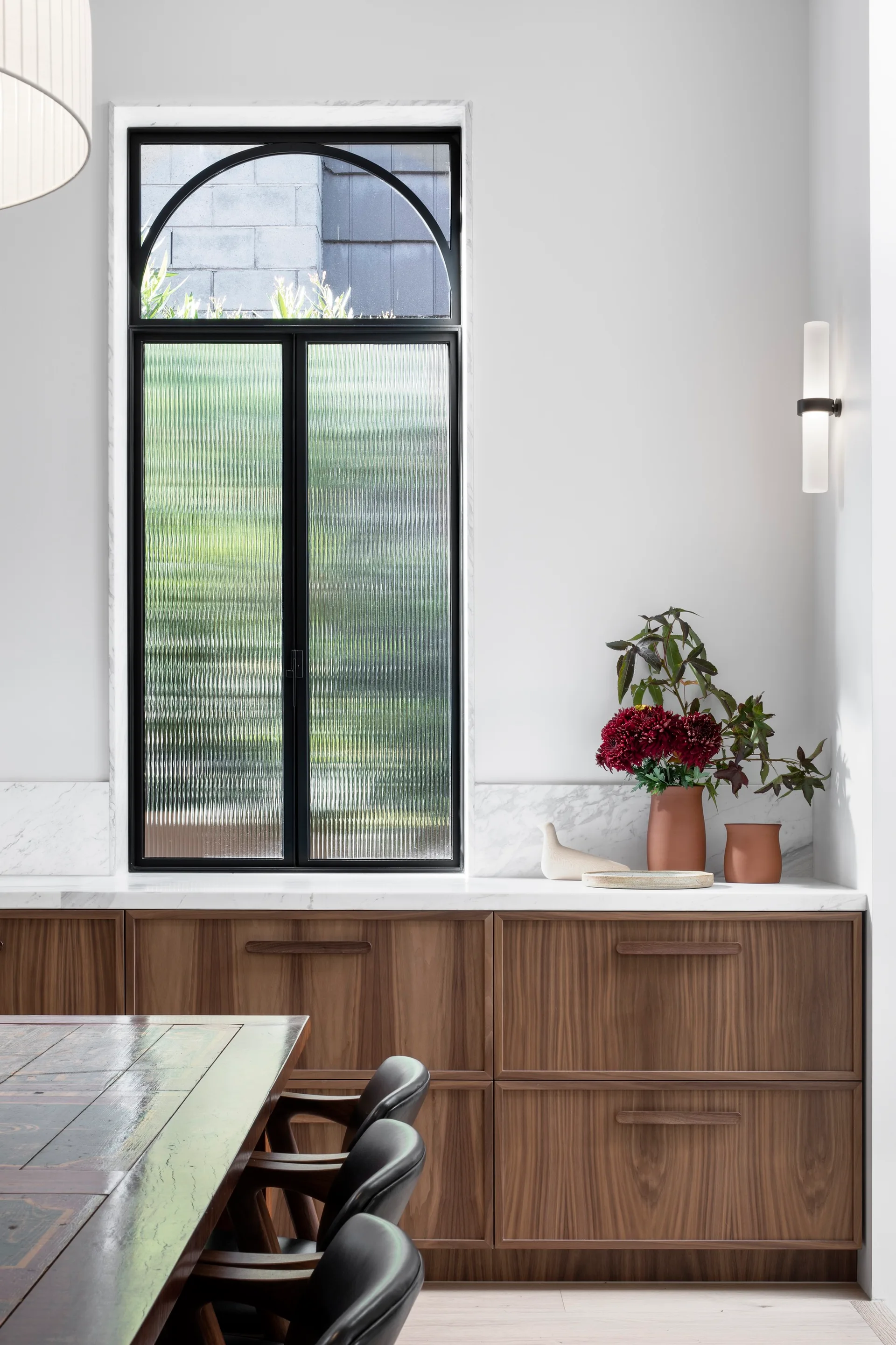 A modern kitchen dining area features wood cabinets with a marble countertop beneath a large arched window with fluted glass.