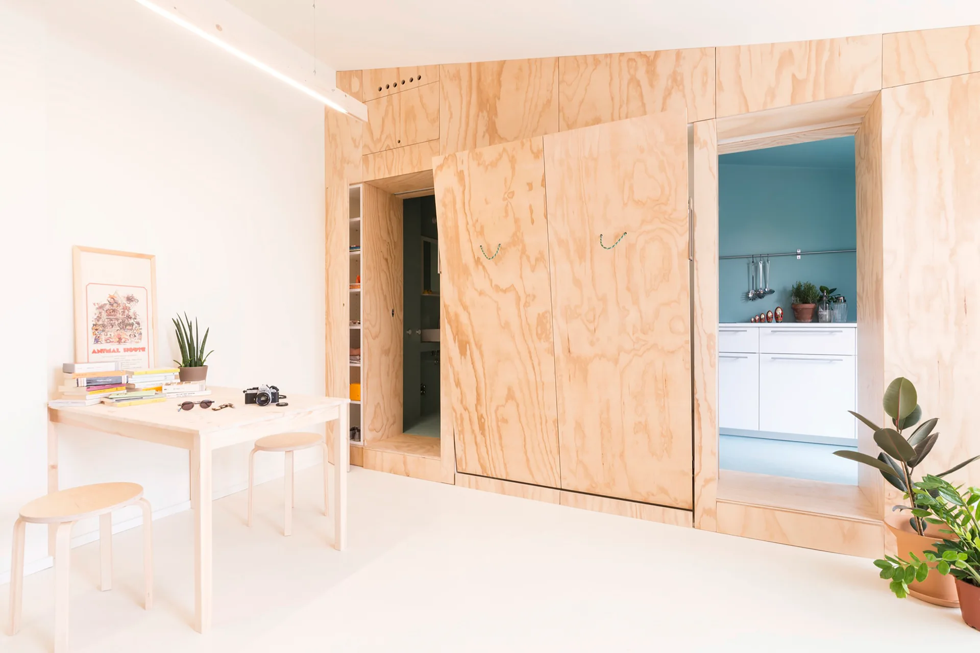 A light wood table and stools sit in a minimalist room next to a large plywood partition wall with doorways opening to a teal kitchen.