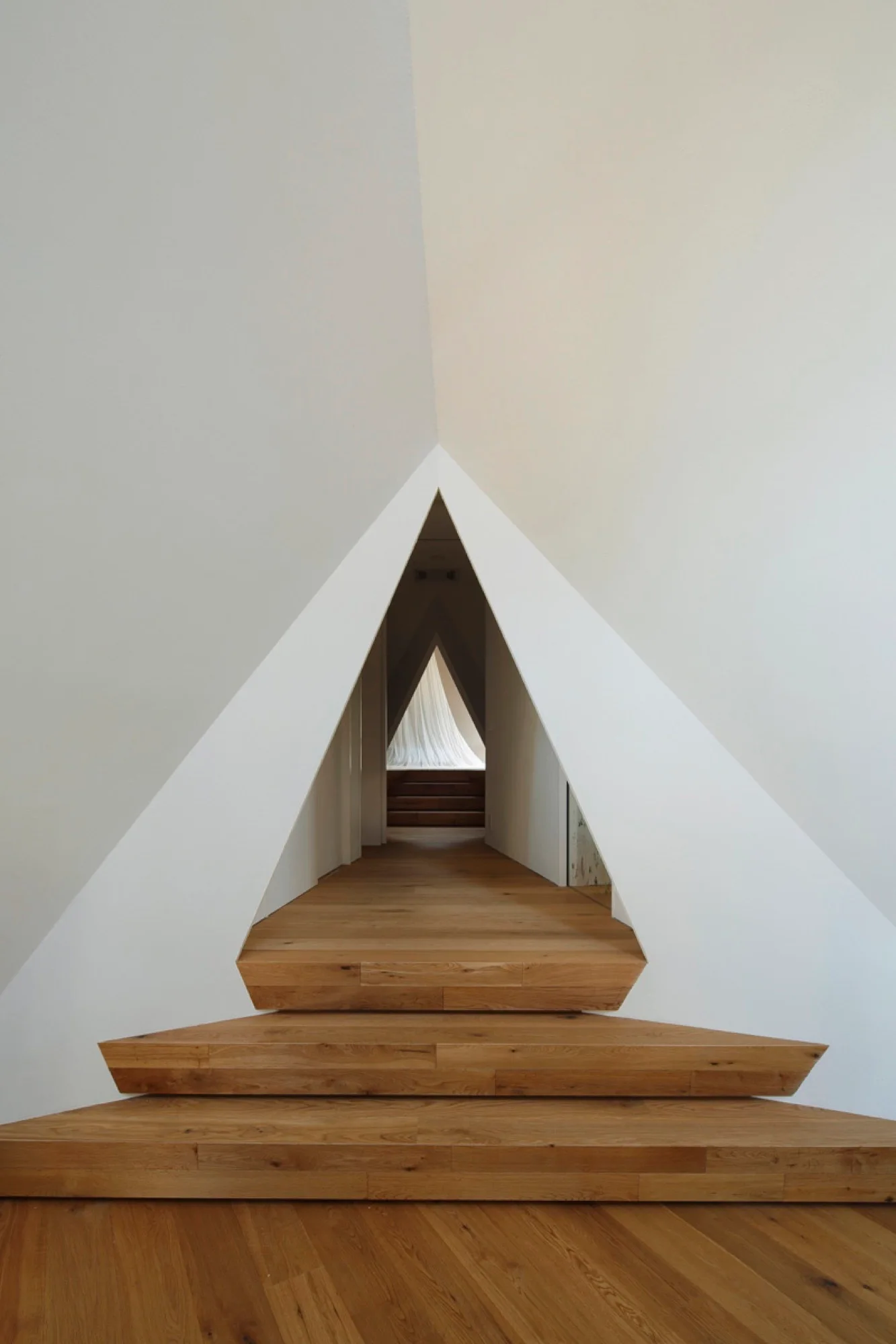 A minimalist white hallway featuring a series of geometric triangular doorways and wide warm wood steps.