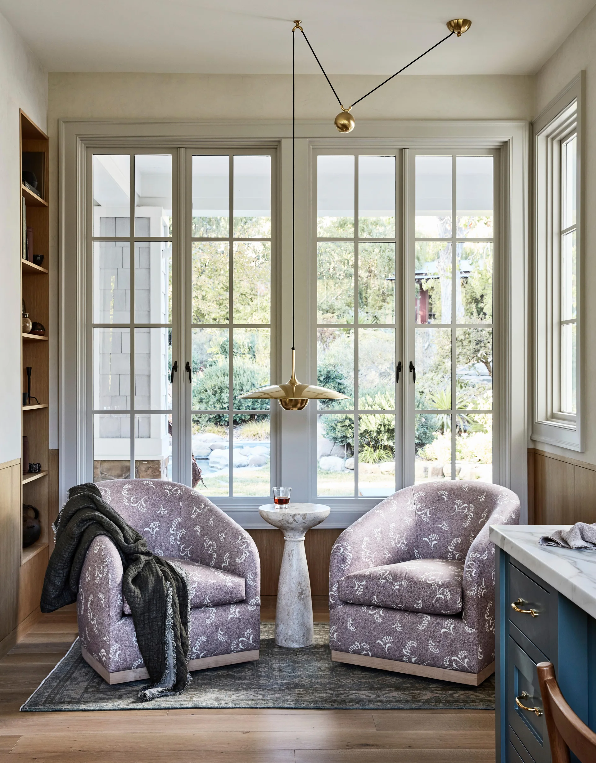 Two mauve patterned armchairs flank a stone pedestal table in front of large multi-pane windows, beneath a hanging brass pendant light.