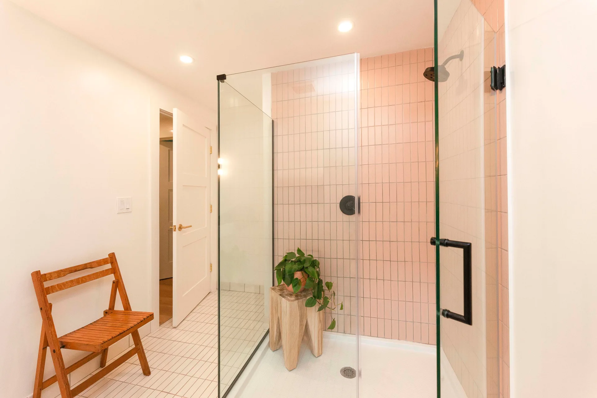 A modern bathroom features a glass-enclosed walk-in shower with pink vertically stacked tiles, matte black fixtures, and a wooden stool.