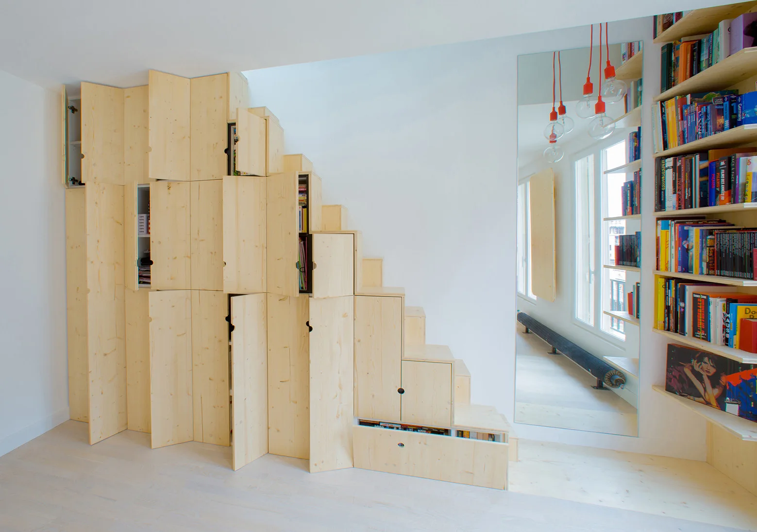 Stepped light wood cabinets built under a staircase stand next to a tall wall mirror reflecting red pendant lights.