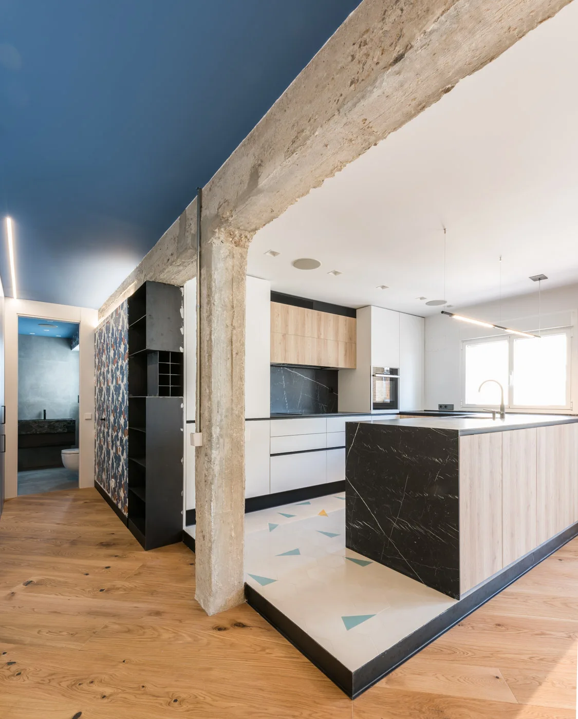 A modern kitchen featuring a dark stone-topped island, light wood cabinets, and a prominent raw concrete support pillar next to geometric tile flooring.