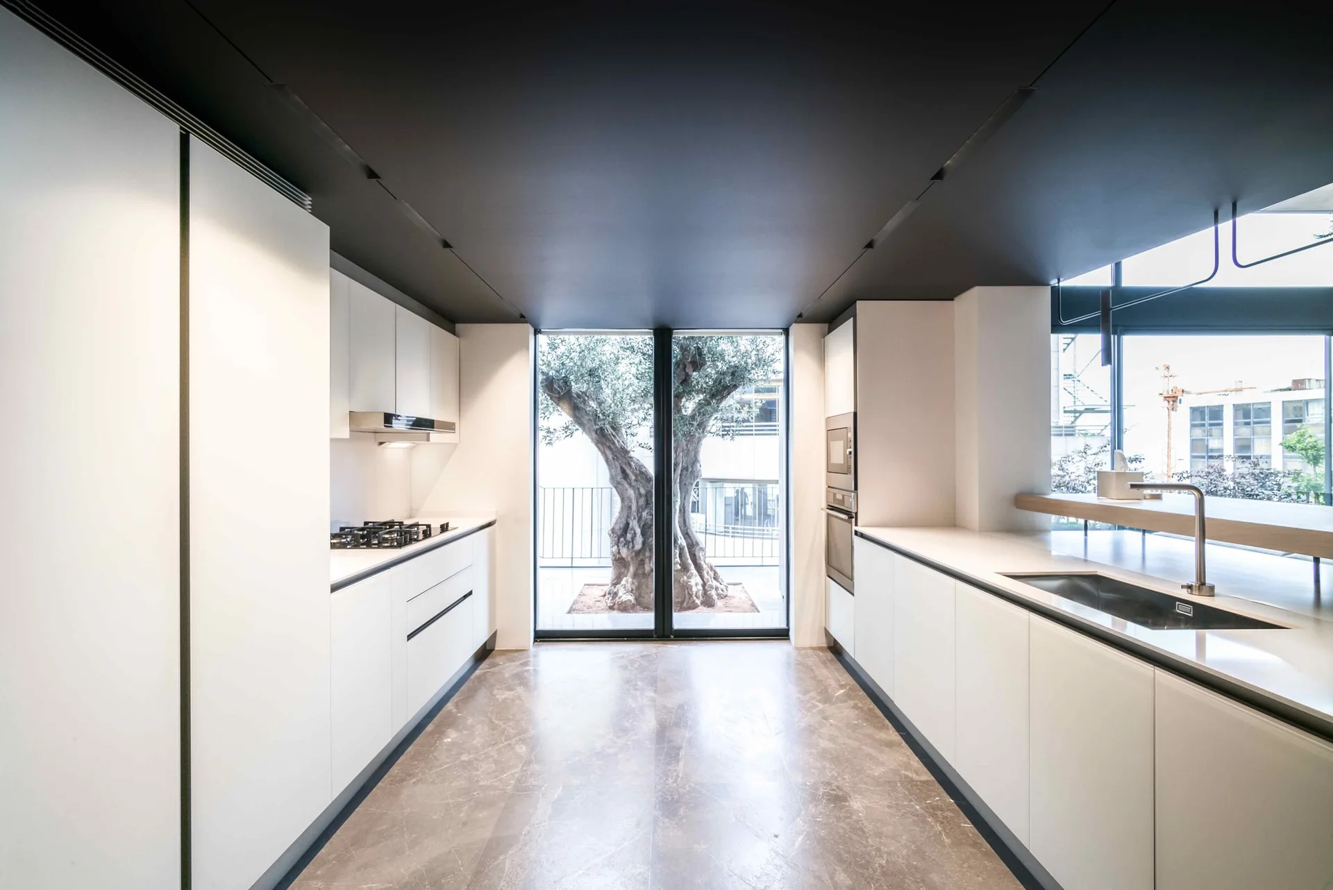 Symmetrical white galley kitchen featuring a charcoal ceiling, grey marbled floor, and a large window looking out at an olive tree.