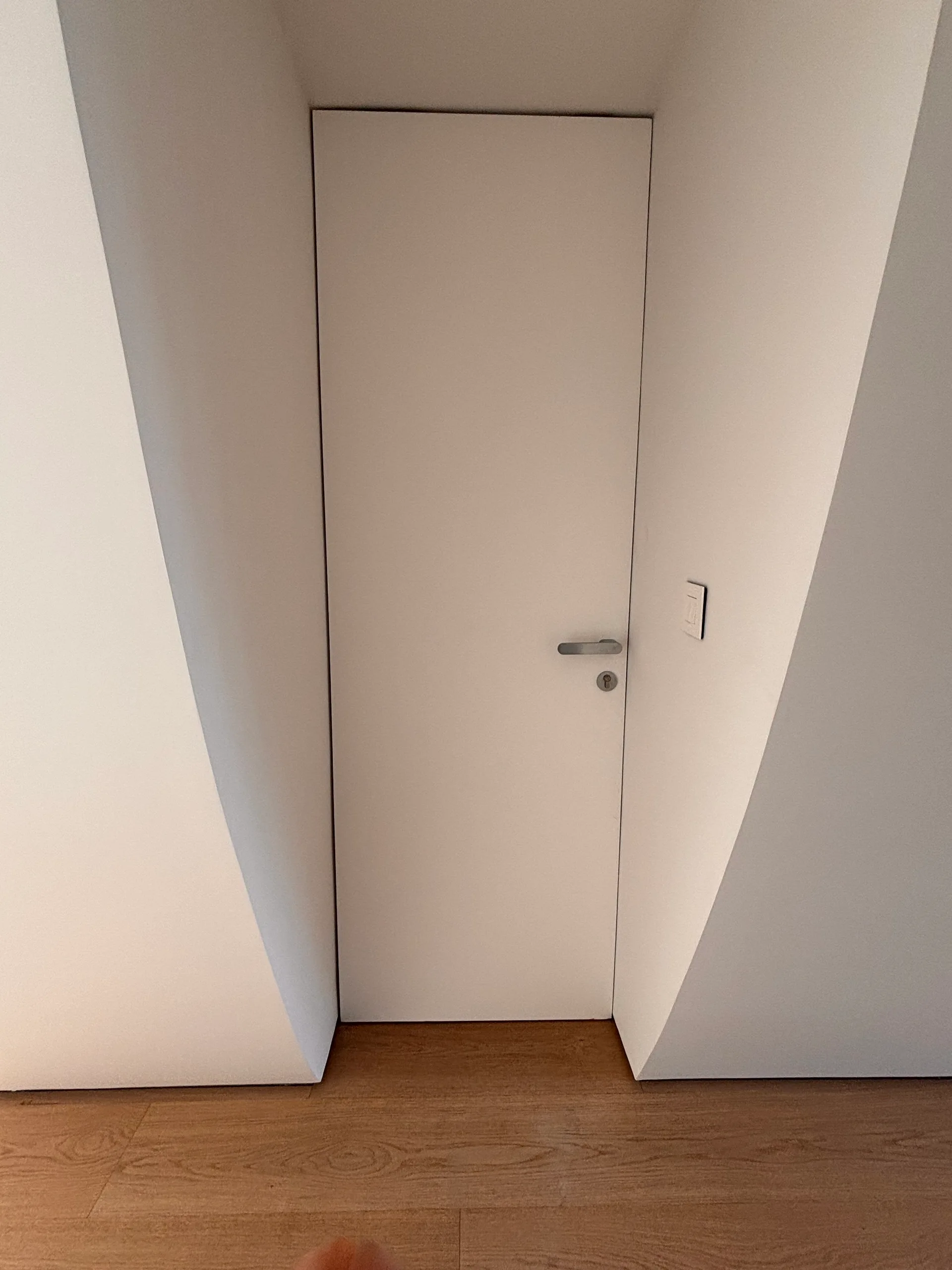 A flush white door is set into a deep recess with slanted white walls above light wood plank flooring.