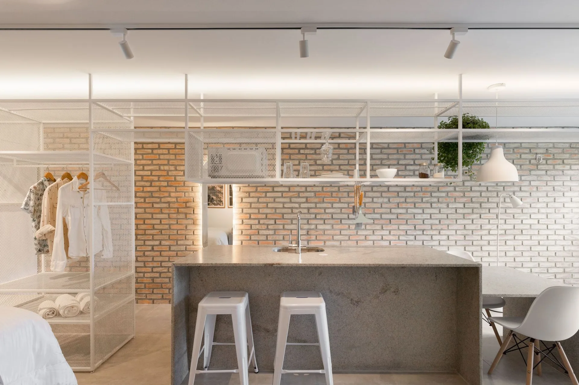 An open-concept kitchen features a textured brick wall, a gray stone island with white stools, and suspended white wire shelving.
