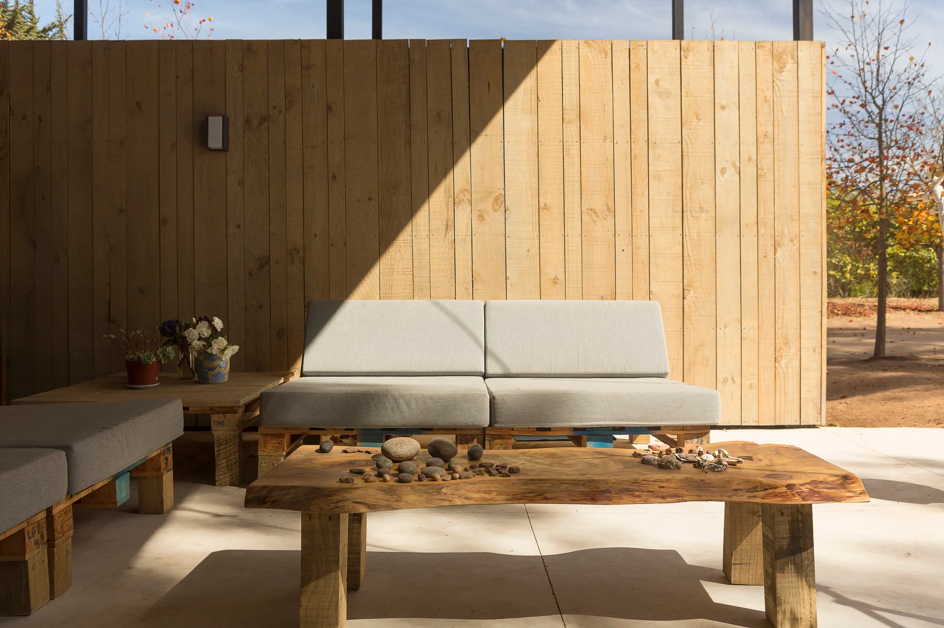 A light gray sofa with a wooden pallet base sits on a concrete patio next to a live edge coffee table and a vertical wood plank wall.
