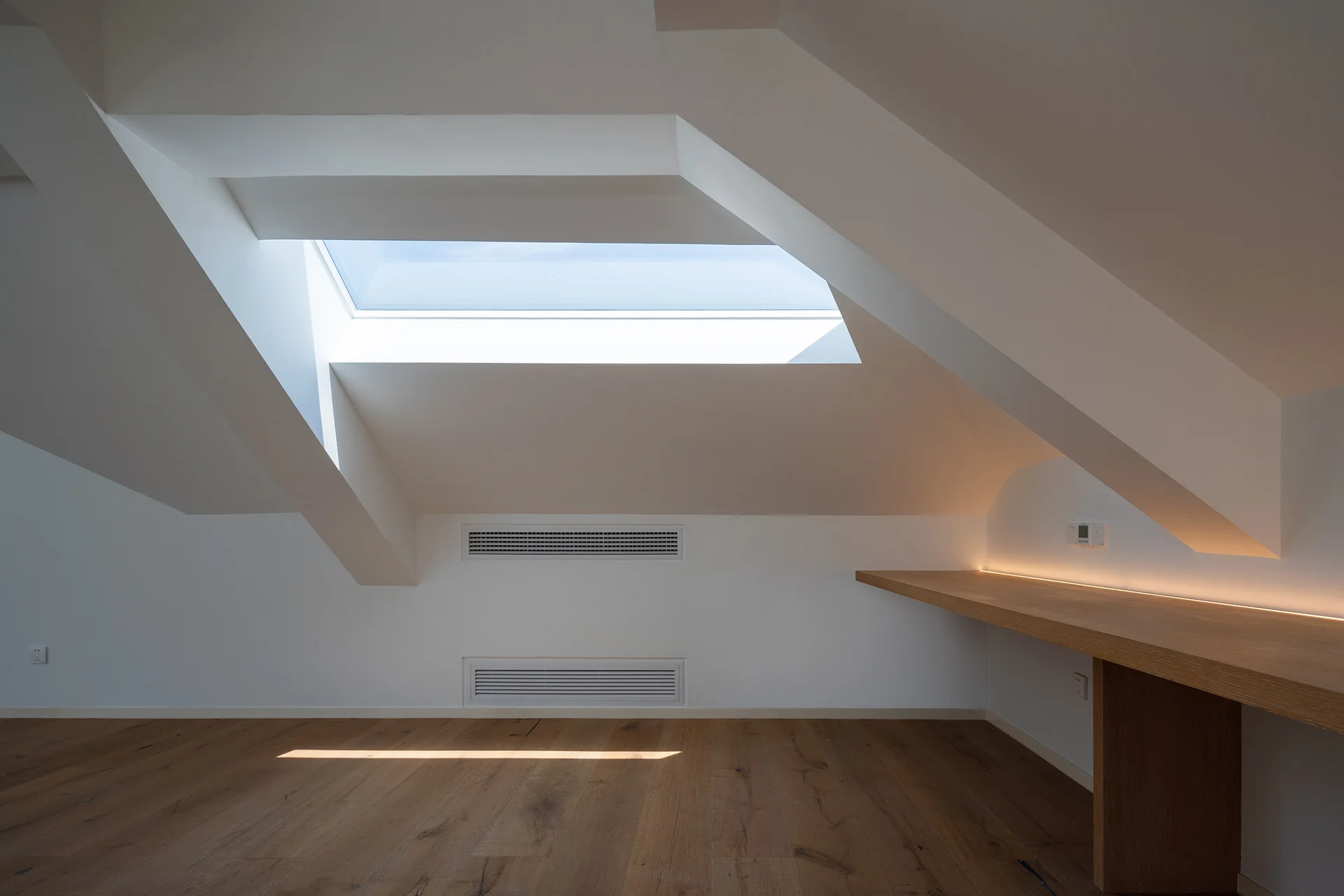 A minimalist attic space featuring a slanted white ceiling with a skylight, a built-in wooden desk with LED backlighting, and wood plank flooring.