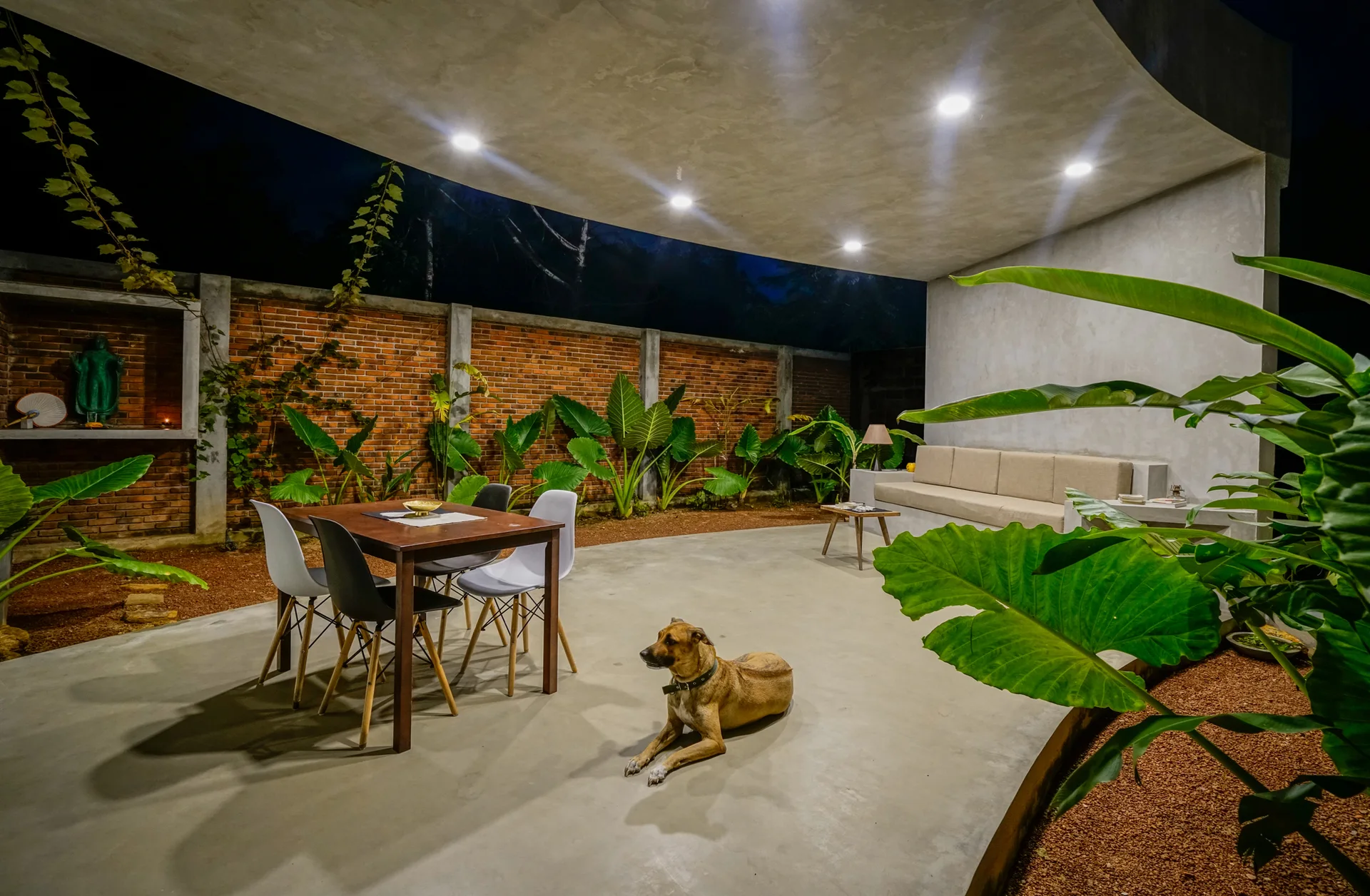 An open-air concrete patio at night features a dining table, a built-in sofa, and large tropical plants against a red brick wall.